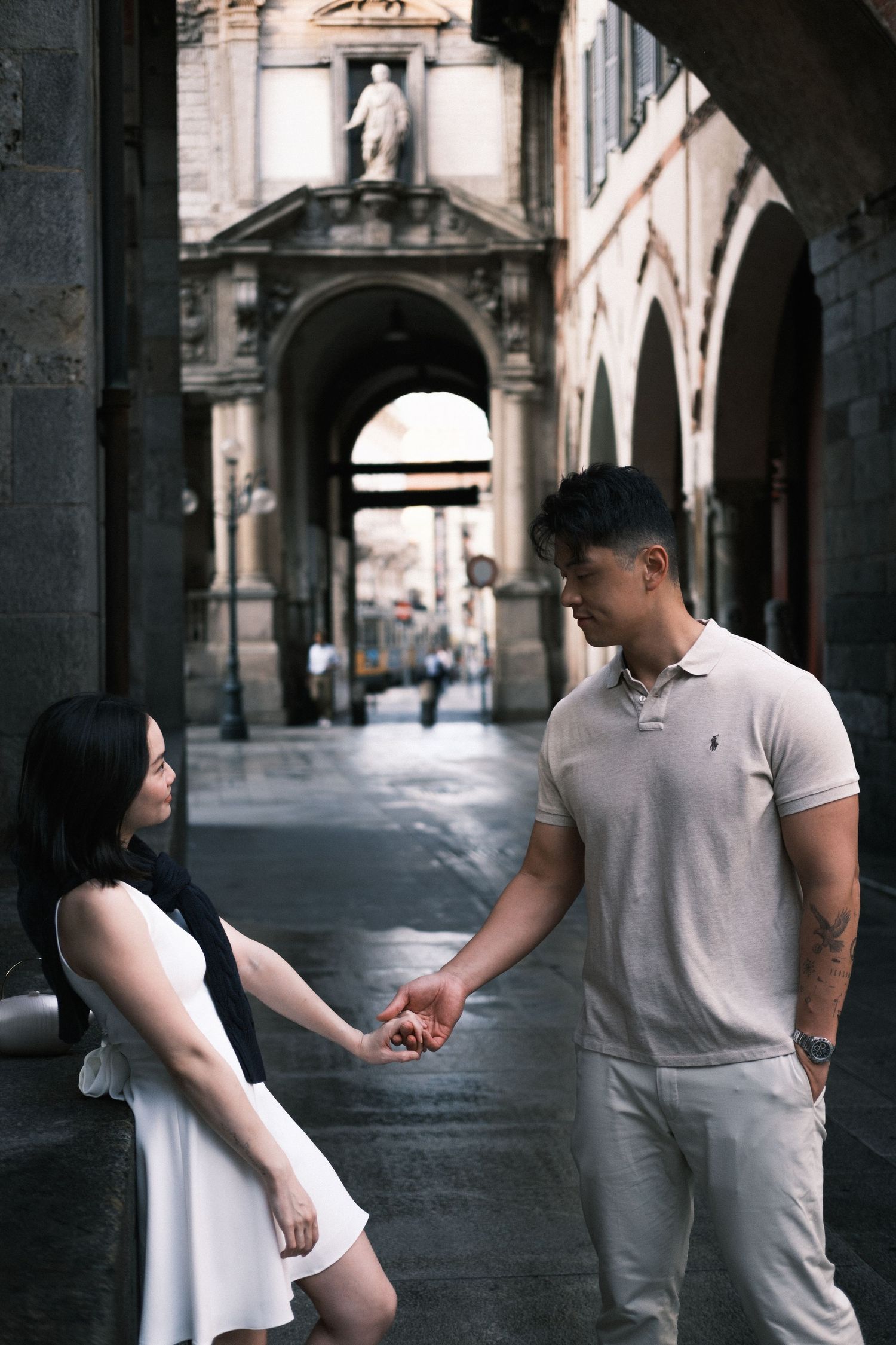 Couple holding hands in a romantic European archway, dressed in casual white outfits on a cobblestone street.
