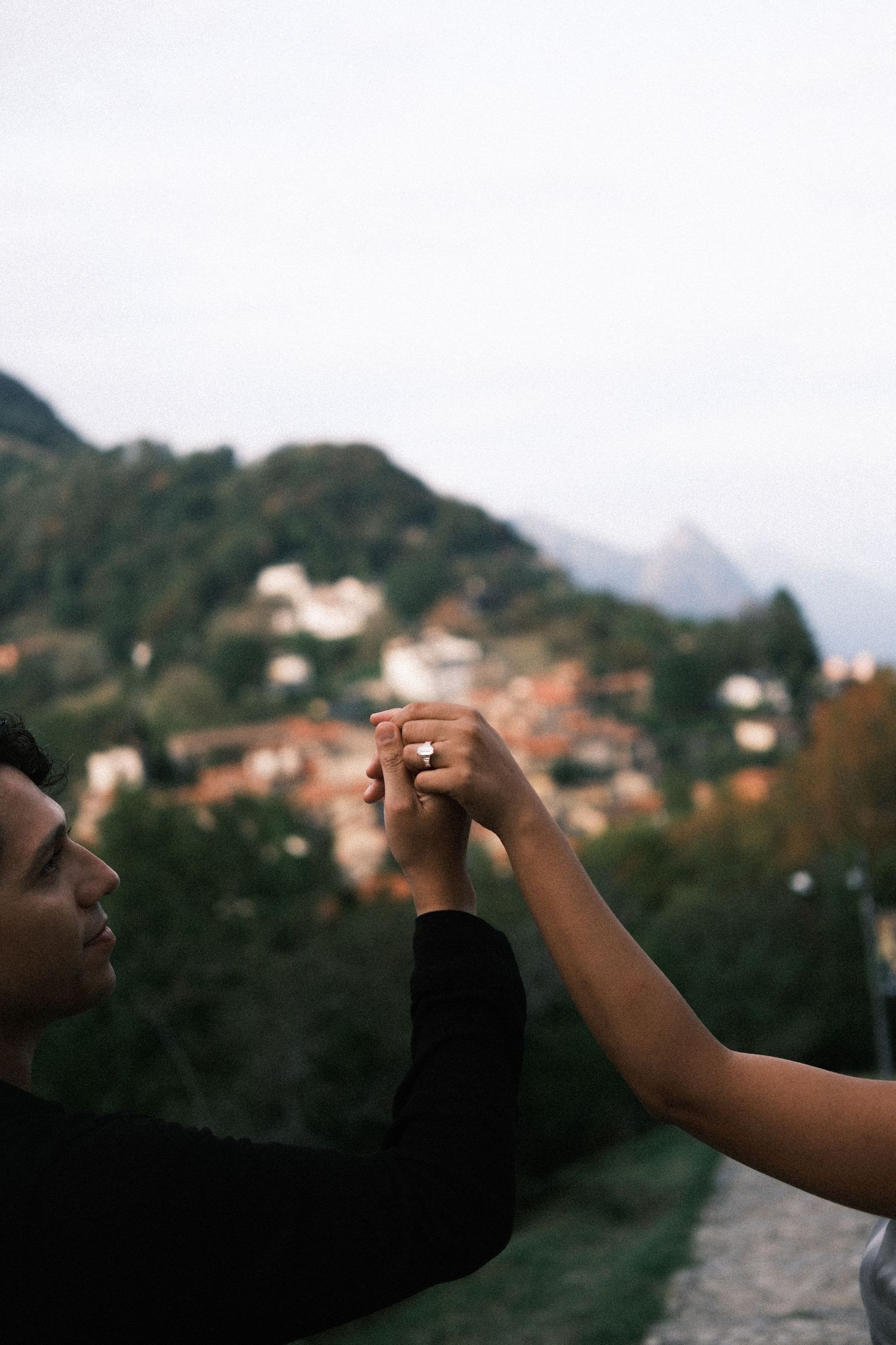 Couple holding hands with engagement ring against scenic mountain village backdrop.