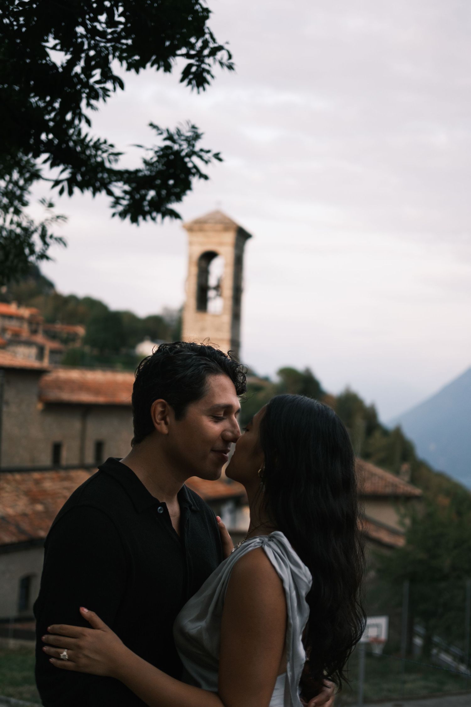 Romantic couple embracing near a historic Italian church bell tower at dusk with mountain views.
