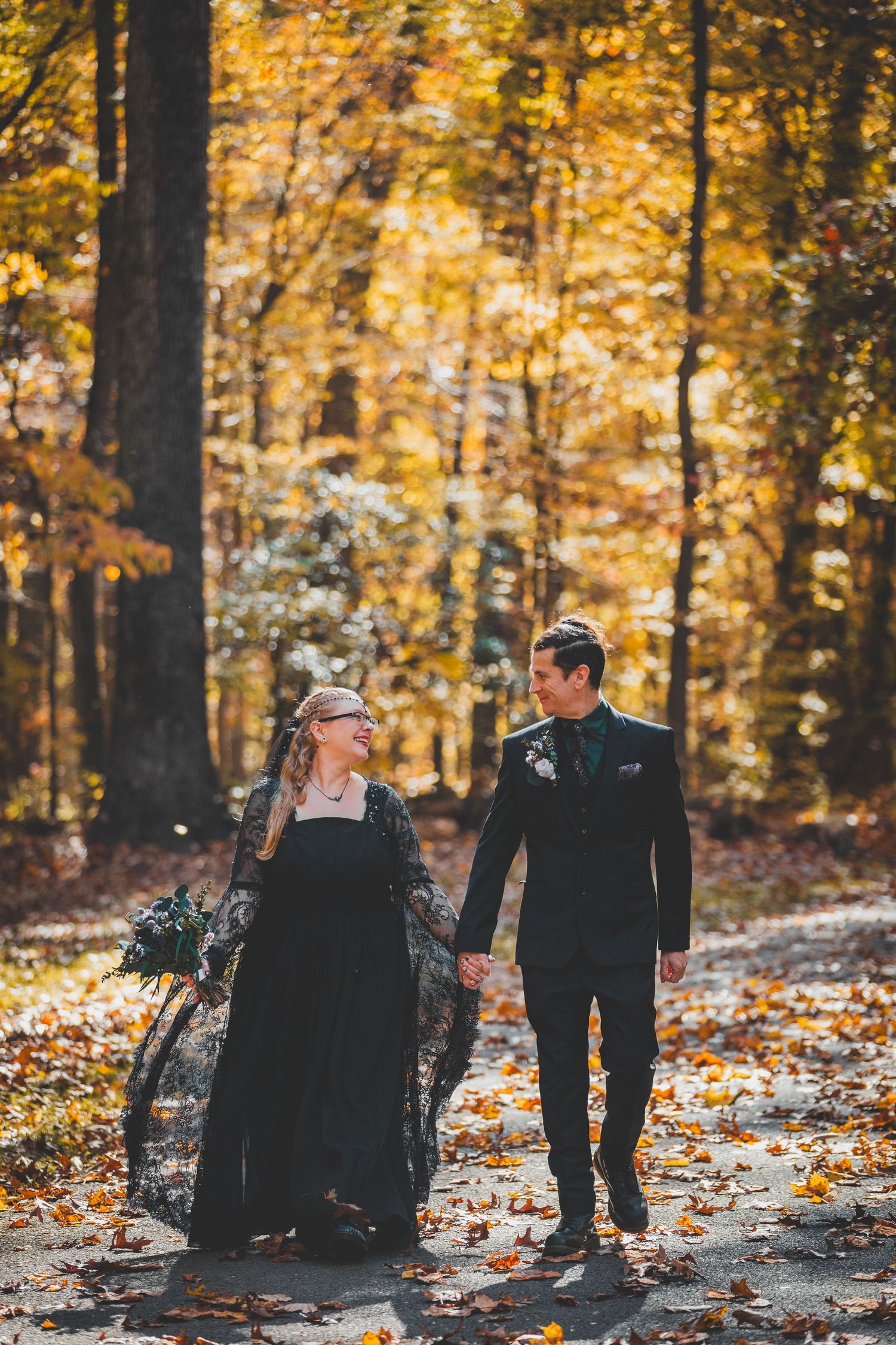Couple in black wedding attire holding hands on a leaf-covered path in a golden autumn forest at Seneca Creek State Park