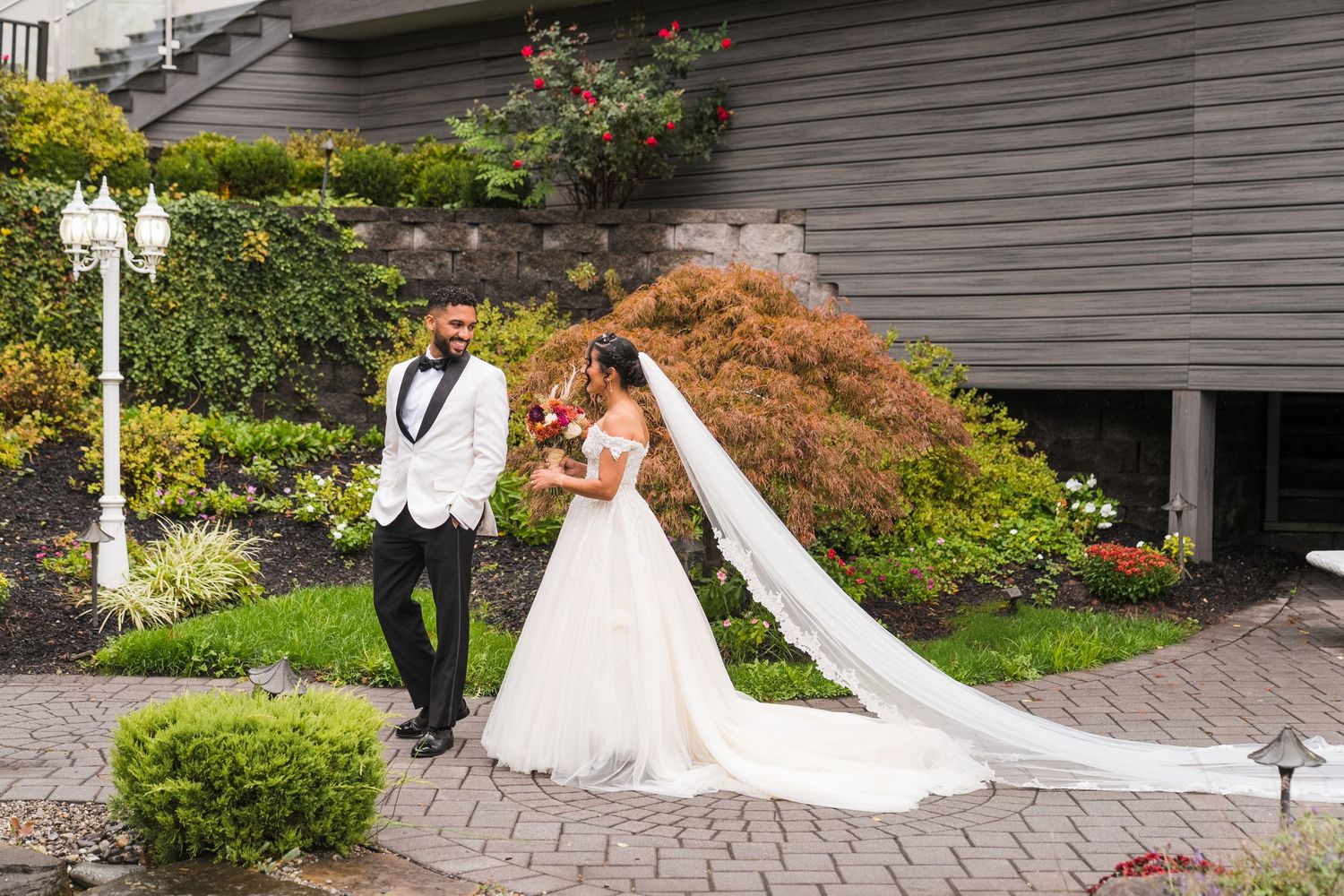 Bride and groom pose in a garden courtyard, her long white veil trailing on stone pavers beside lush greenery.
