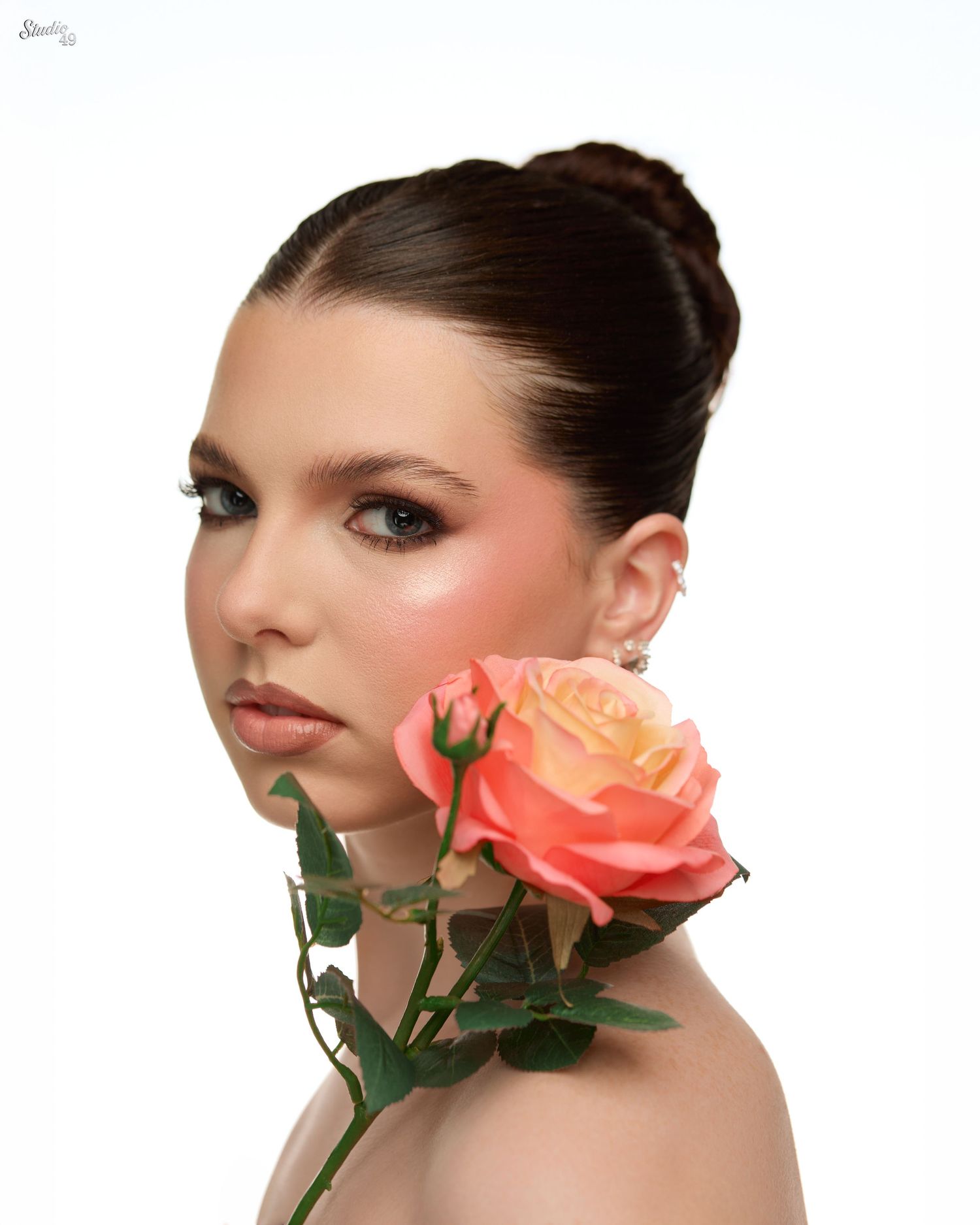 Close-up portrait of young woman with updo hairstyle holding a peach rose near her face.