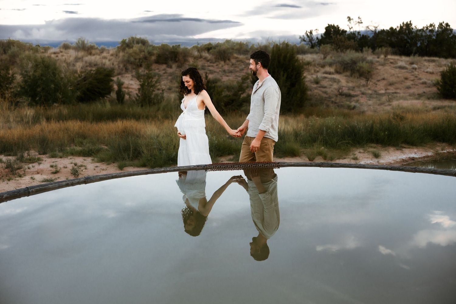 Pregnant couple holds hands walking beside reflective pool in desert landscape, woman in white dress, maternity photo.
