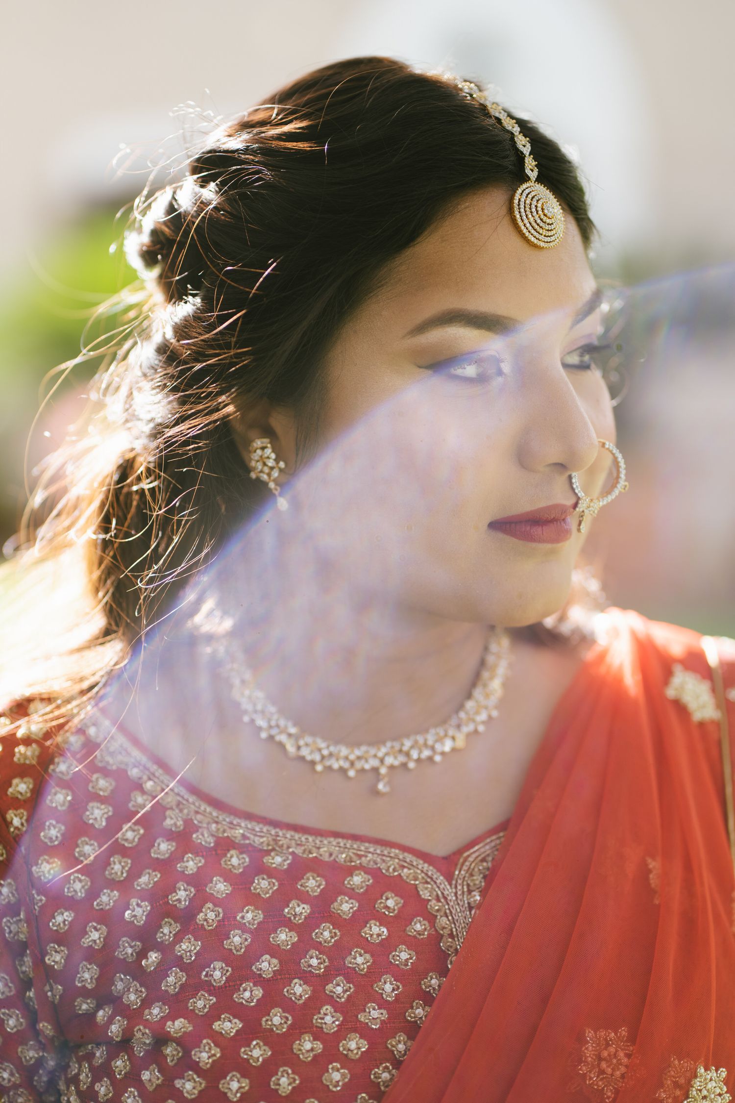 Indian bride in red lehenga with gold maang tikka, nose ring, and diamond necklace, posing in warm sunlight.