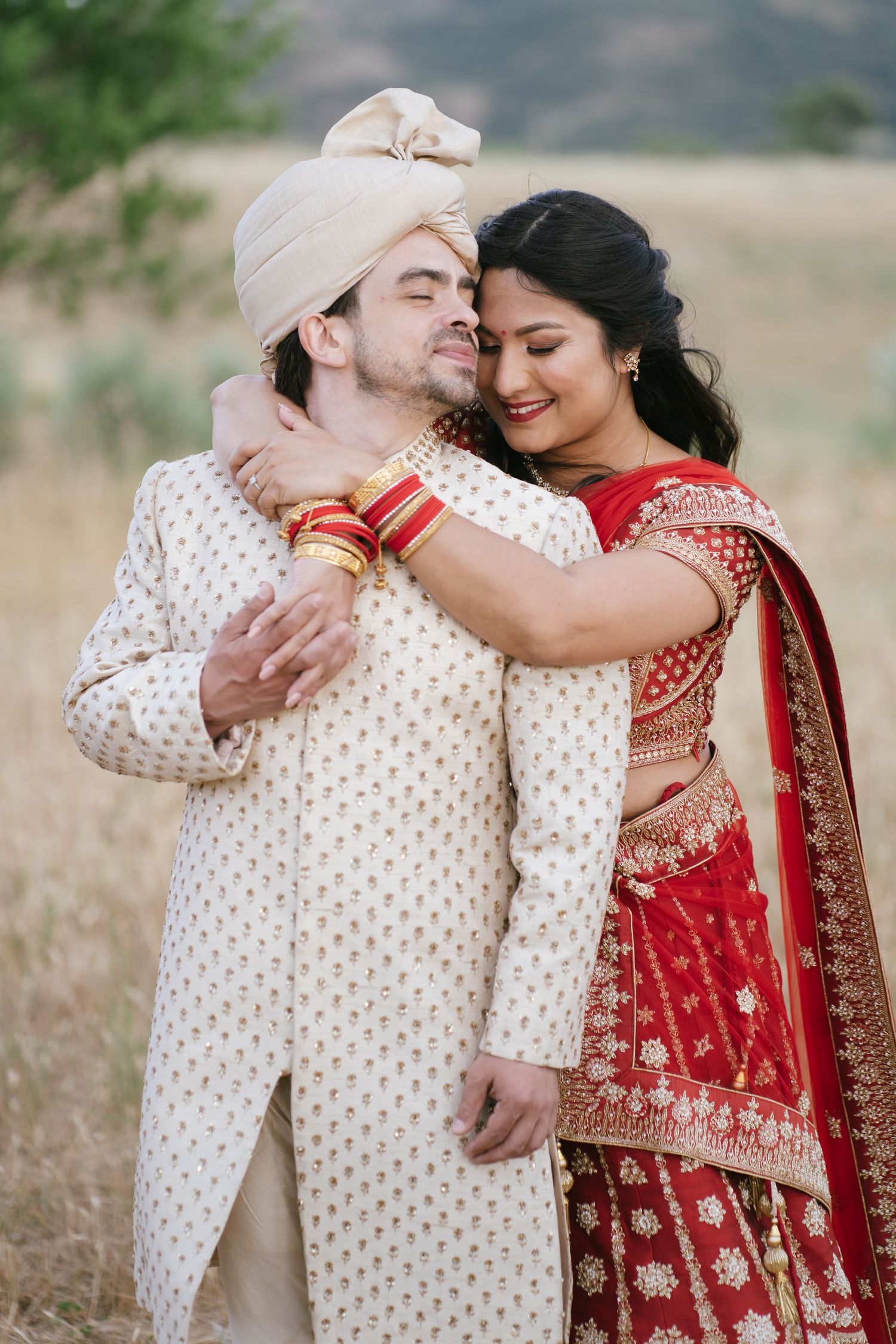 Indian bride in red lehenga embraces groom in cream sherwani and turban in a golden field.