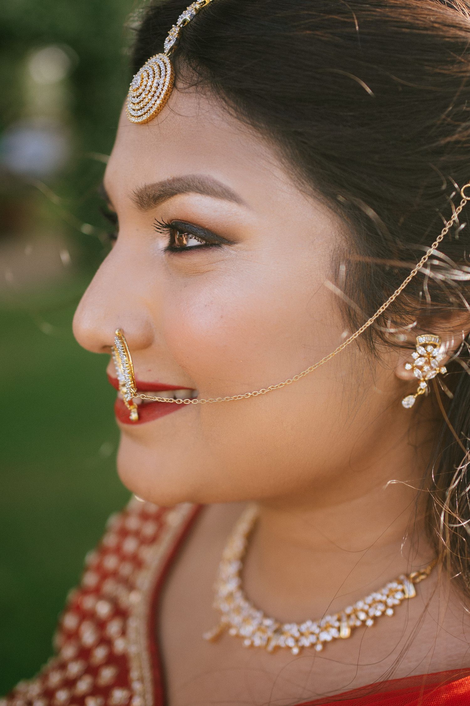 Close-up profile of Indian bride wearing gold maang tikka, nose ring, pearl necklace, and red bridal outfit.