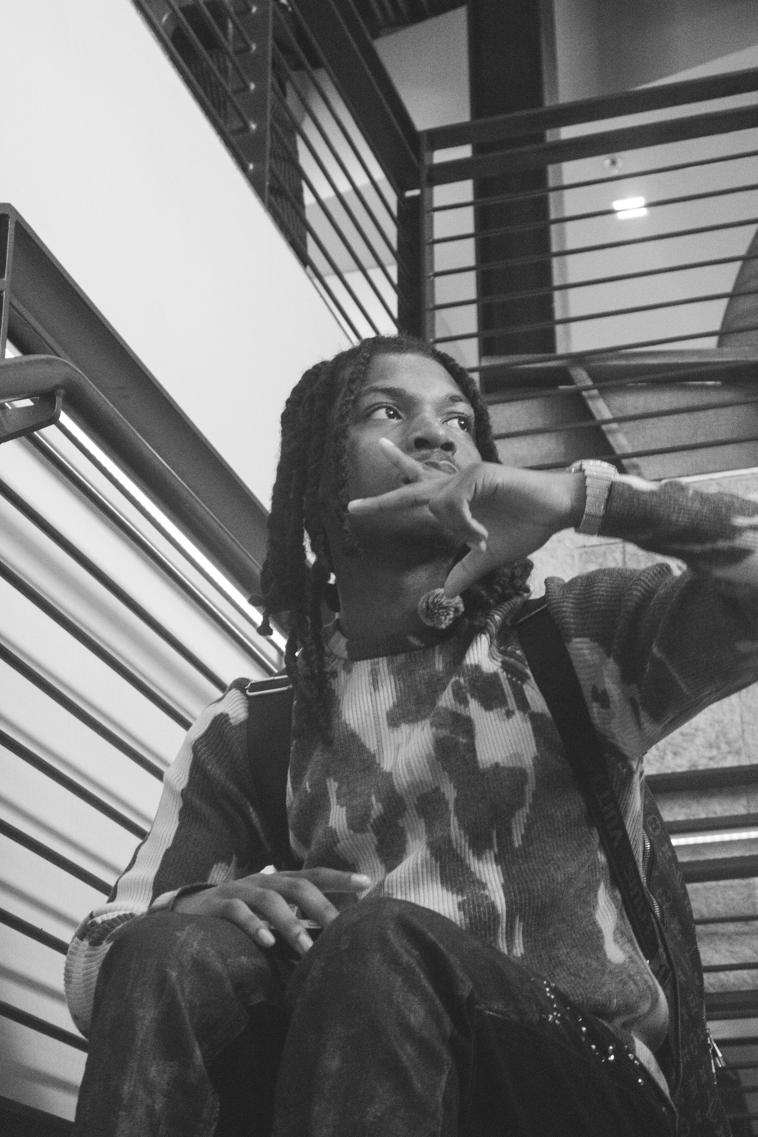 Black and white photo of a young man with dreadlocks sitting on stairs, wearing a patterned sweater, looking away thoughtfully.