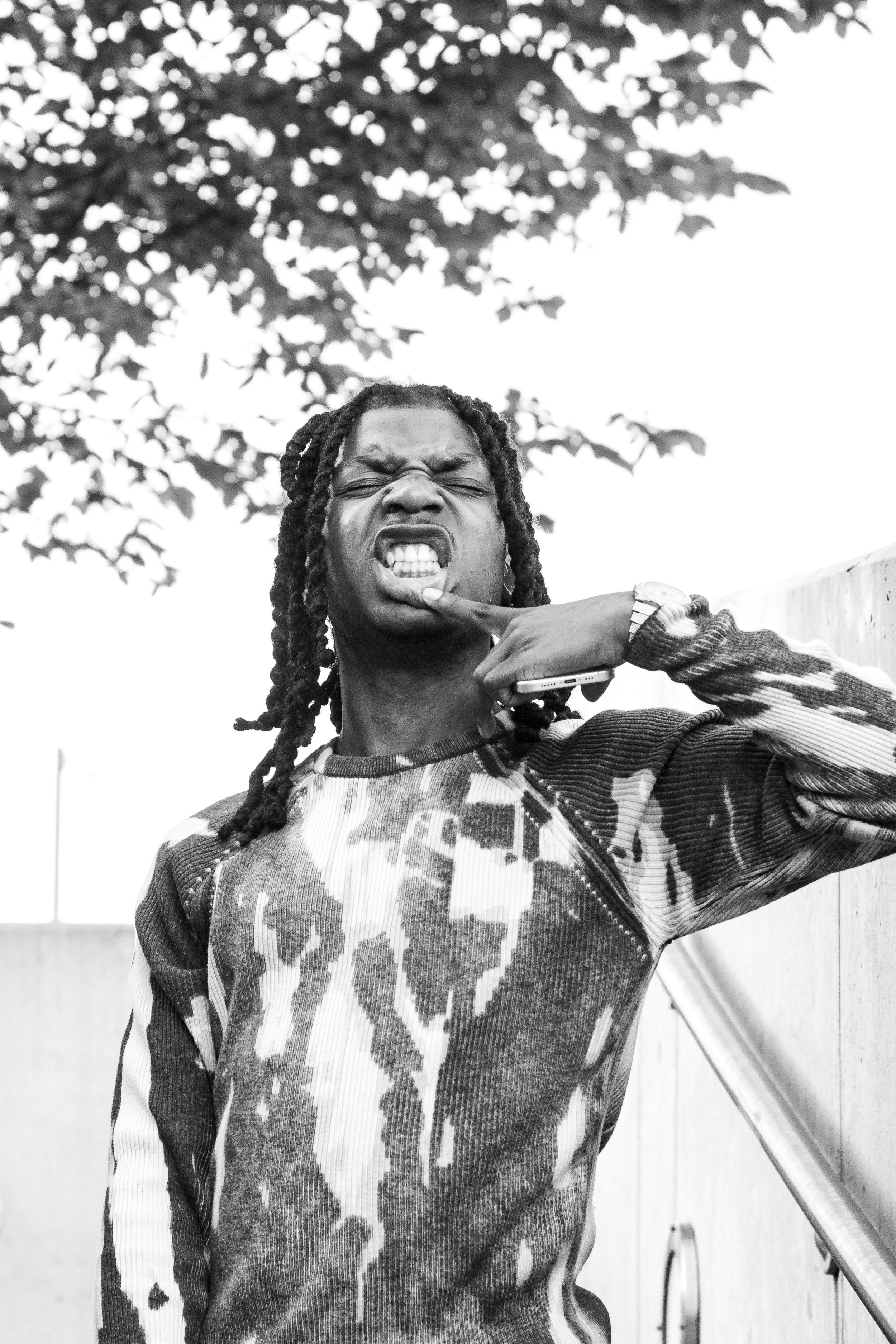 Black and white portrait of a man with dreadlocks in a tie-dye shirt making an expressive face outdoors.