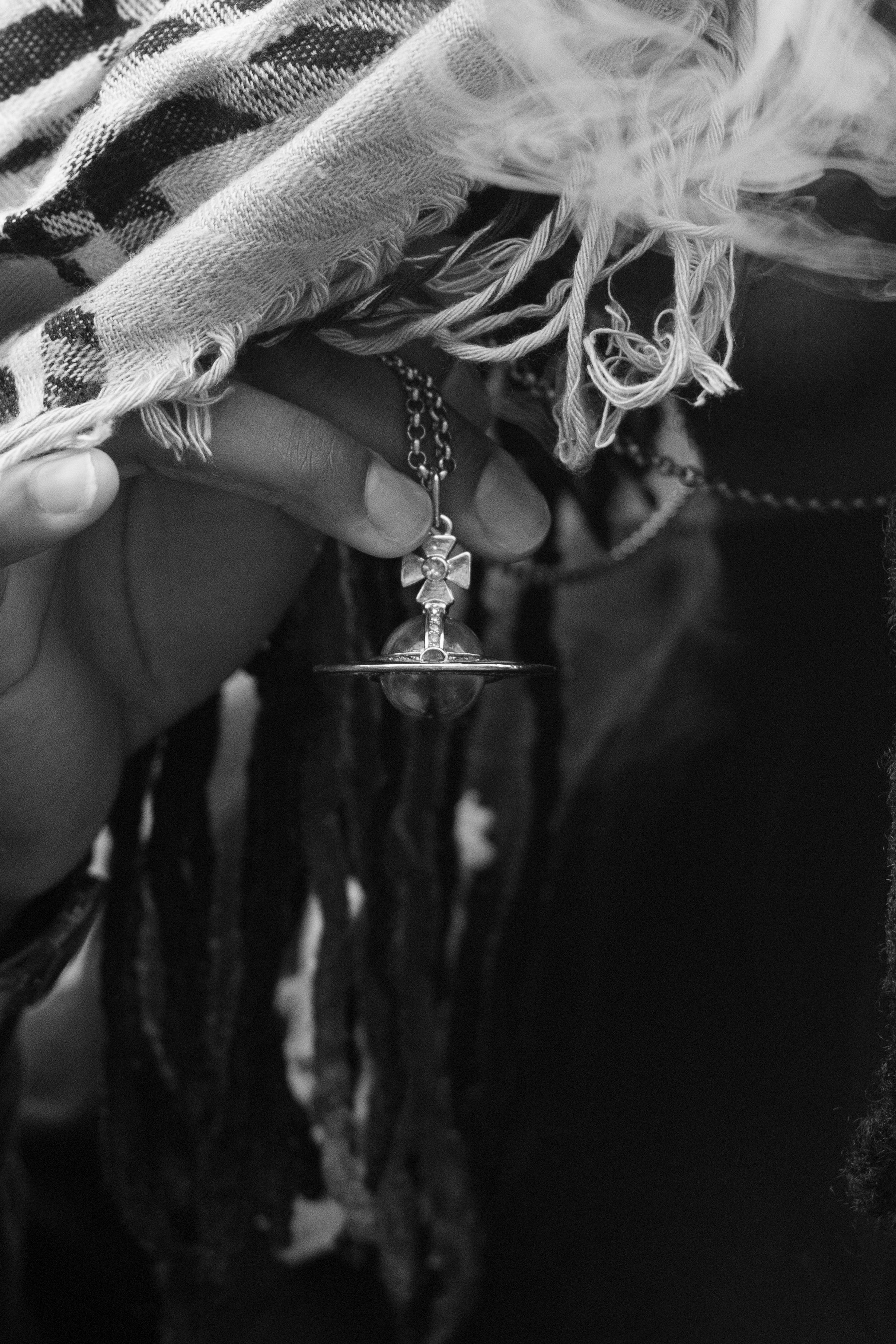 Black and white photo of a hand holding a cross pendant necklace with dreadlocks and fringe scarf in background.
