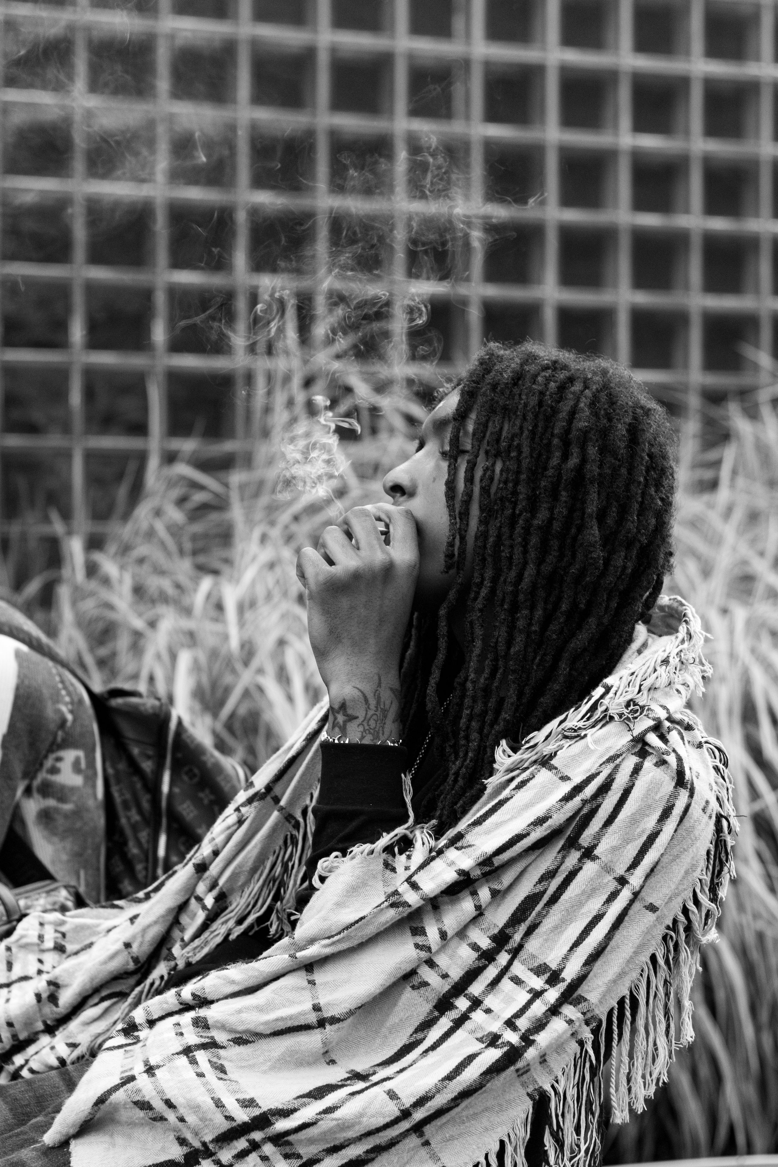 Black and white portrait of a person with dreadlocks wearing a plaid shawl, exhaling smoke outdoors.