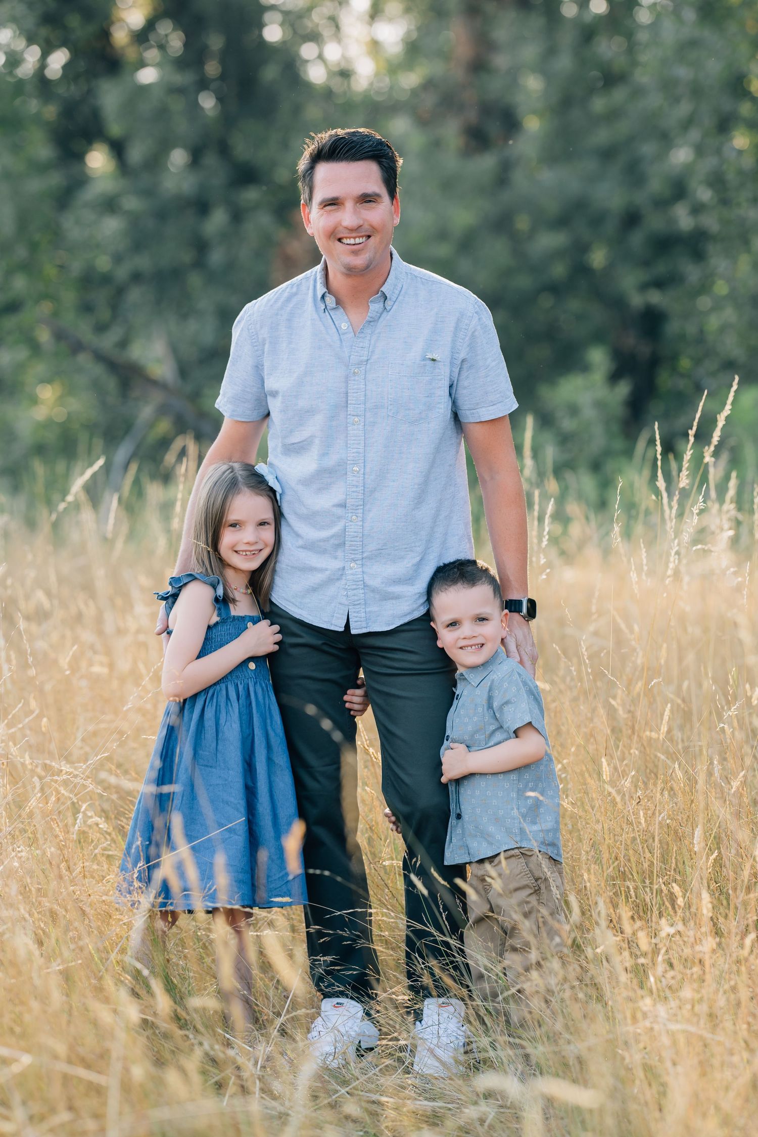 A smiling man poses outdoors in a field with two young children in matching blue outfits, surrounded by tall grass and trees.