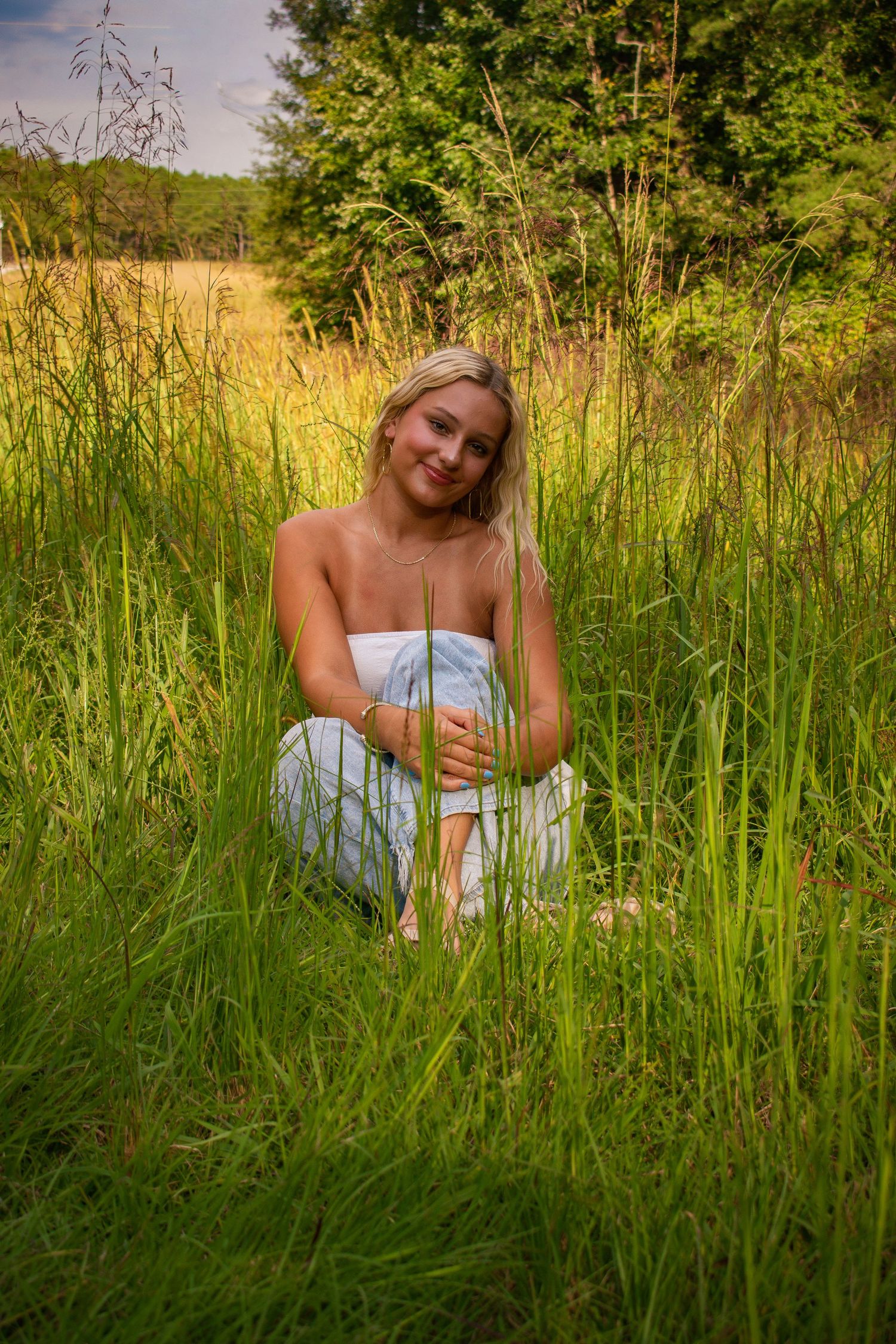 Blonde woman in a white dress crouching and smiling in tall green meadow grass.