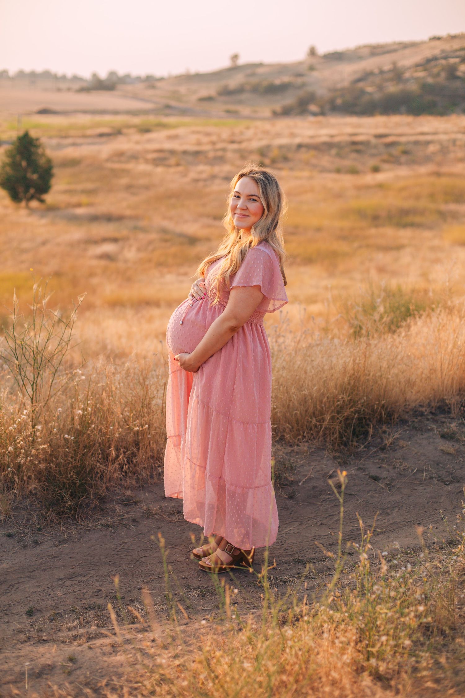 Pregnant woman in pink maxi dress cradling baby bump in golden field at sunset.