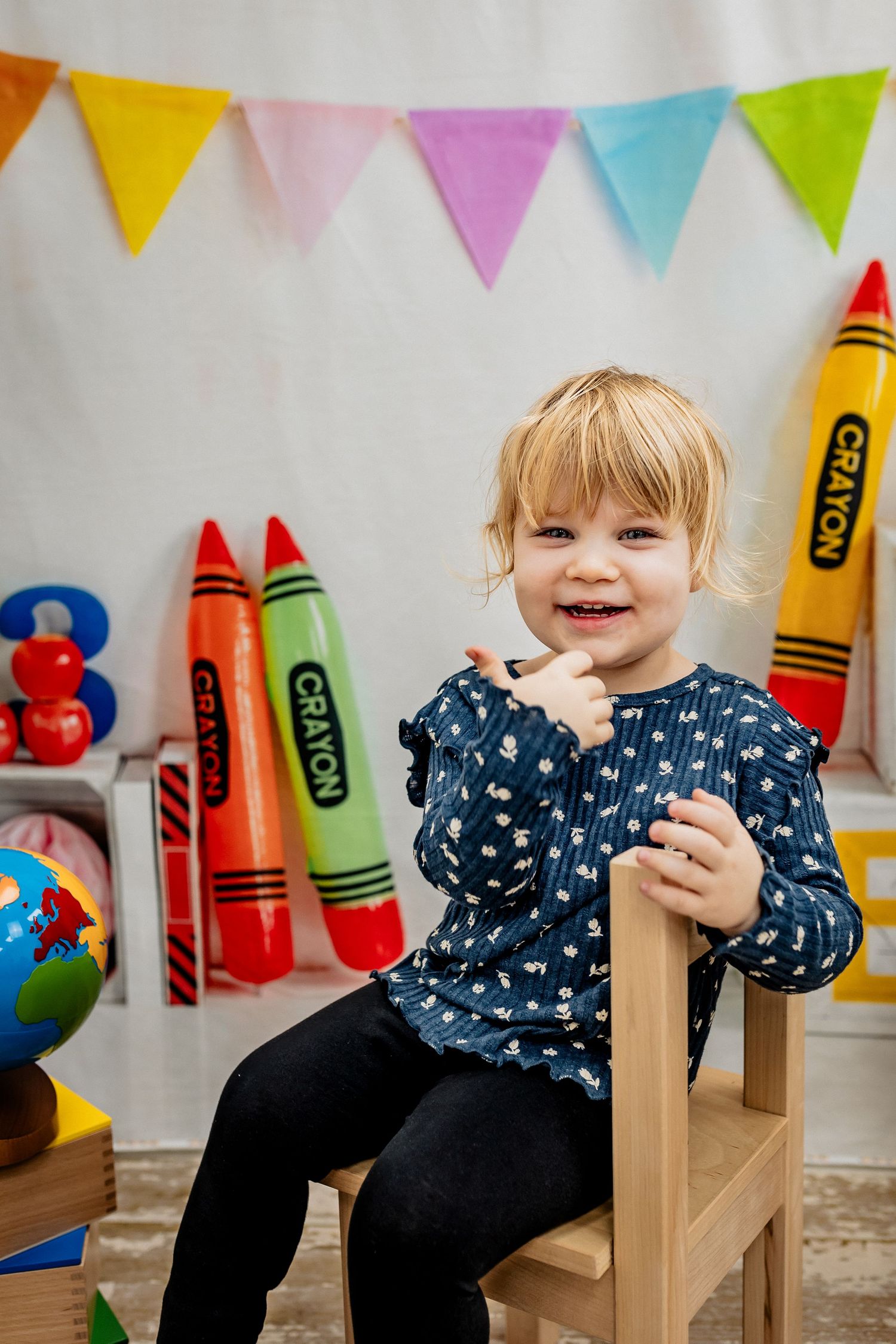 Smiling toddler girl sitting on wooden chair surrounded by colorful crayon decorations and toys in a playful setting.