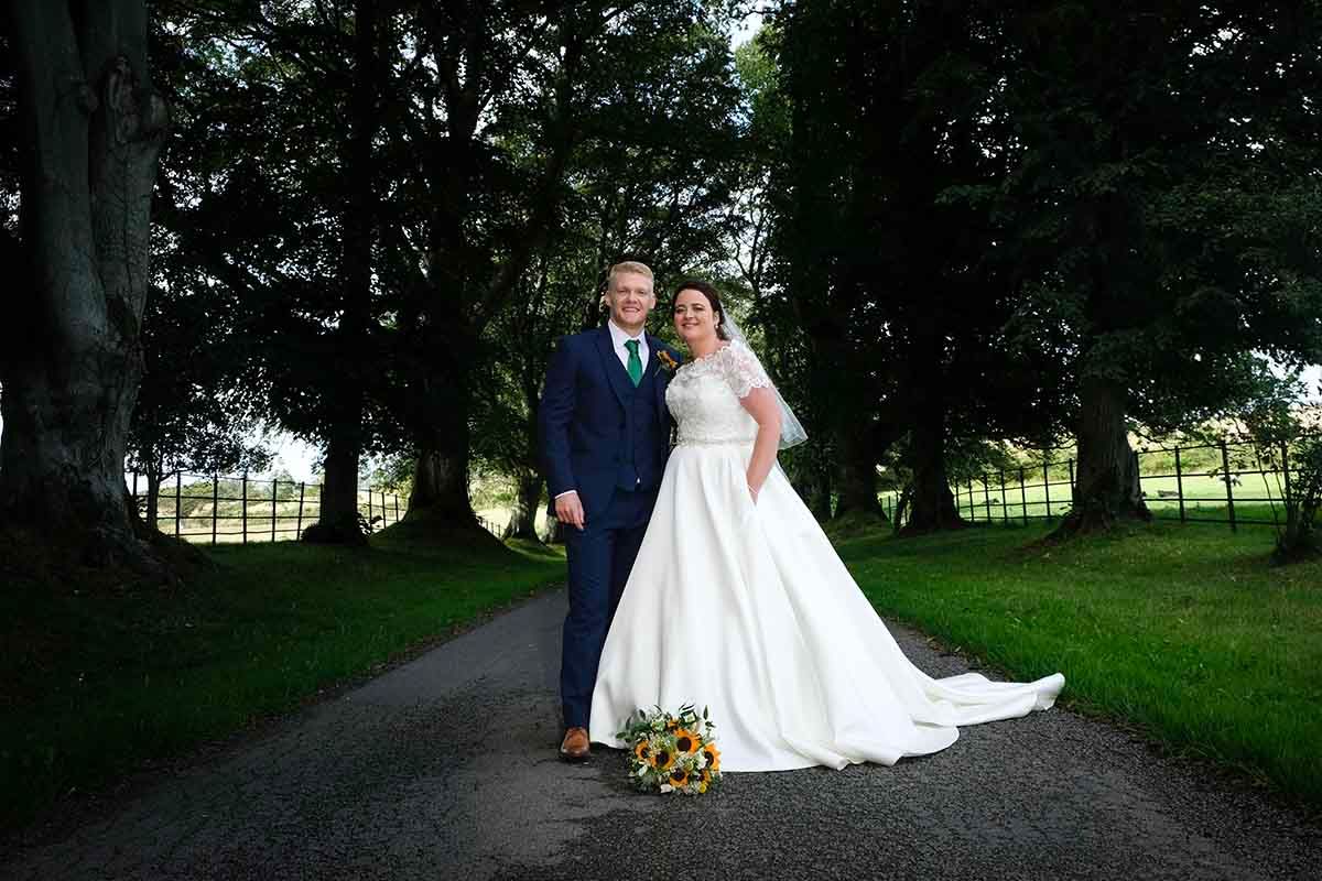 Bride in white ball gown and groom in navy suit pose on a tree-lined driveway at Blaithwaite House wedding venue