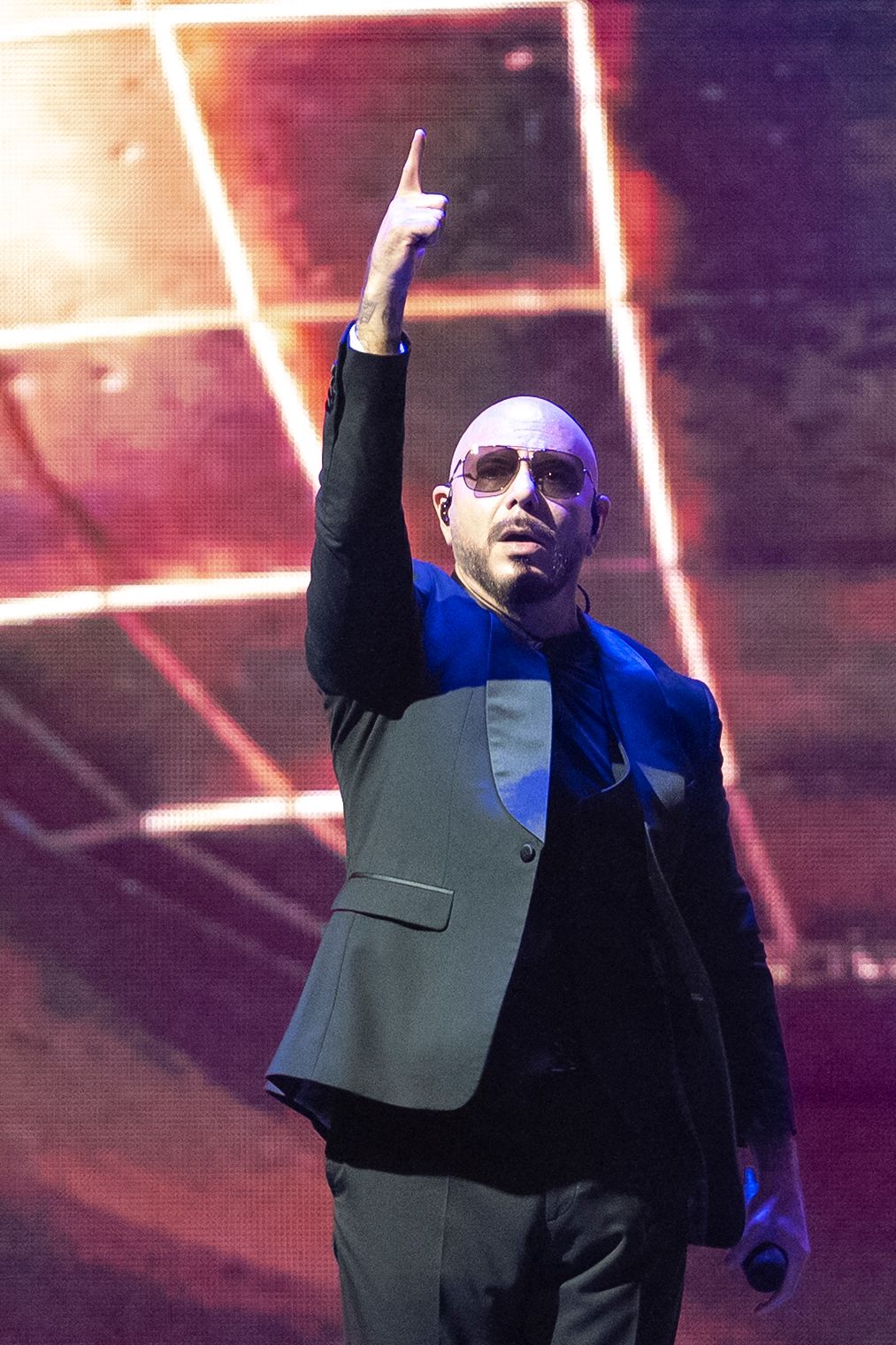 Bald performer in sunglasses and blazer raises index finger on stage with colorful LED backdrop.