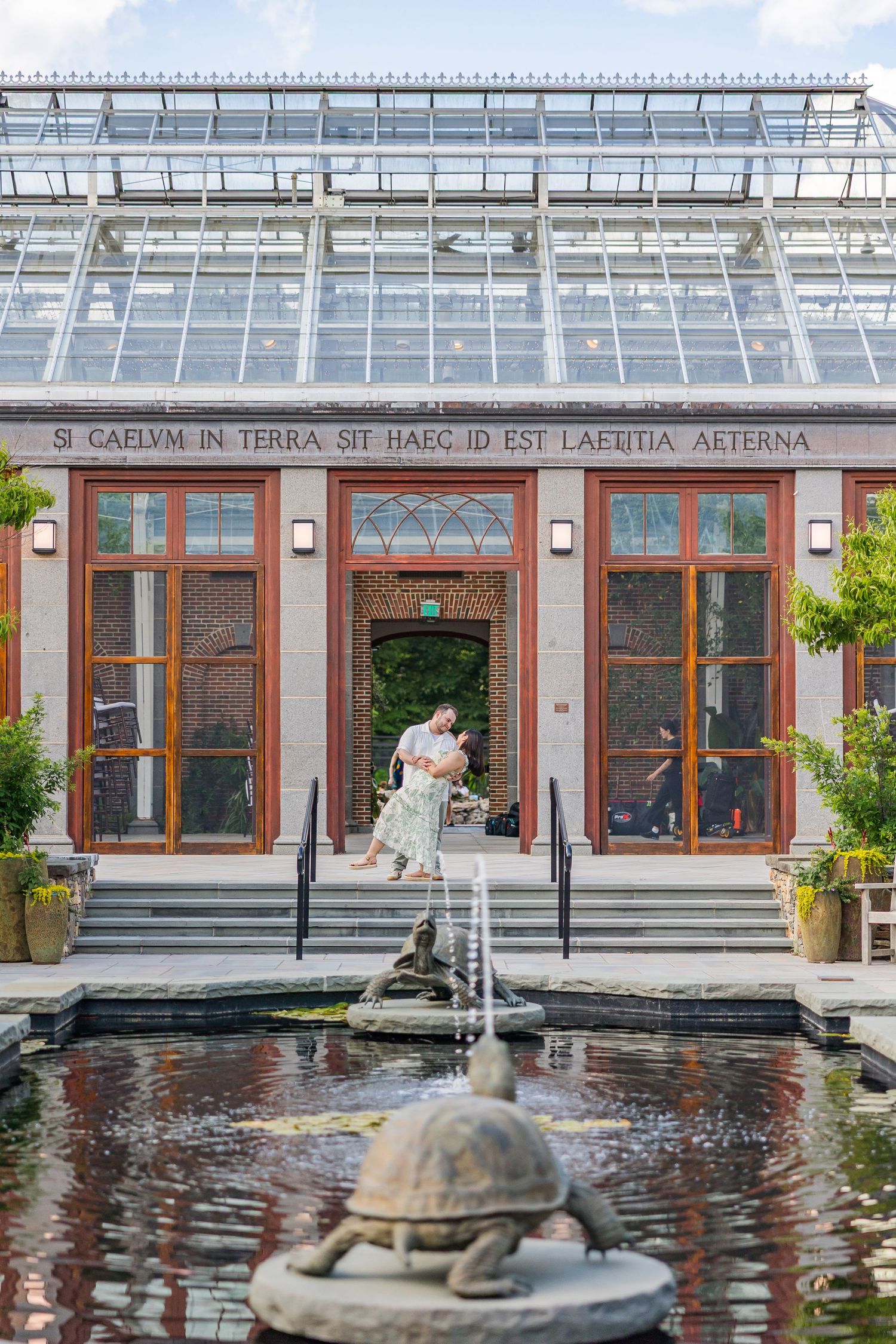 Couple embracing on steps of a glass greenhouse conservatory with fountain and Latin inscription above.