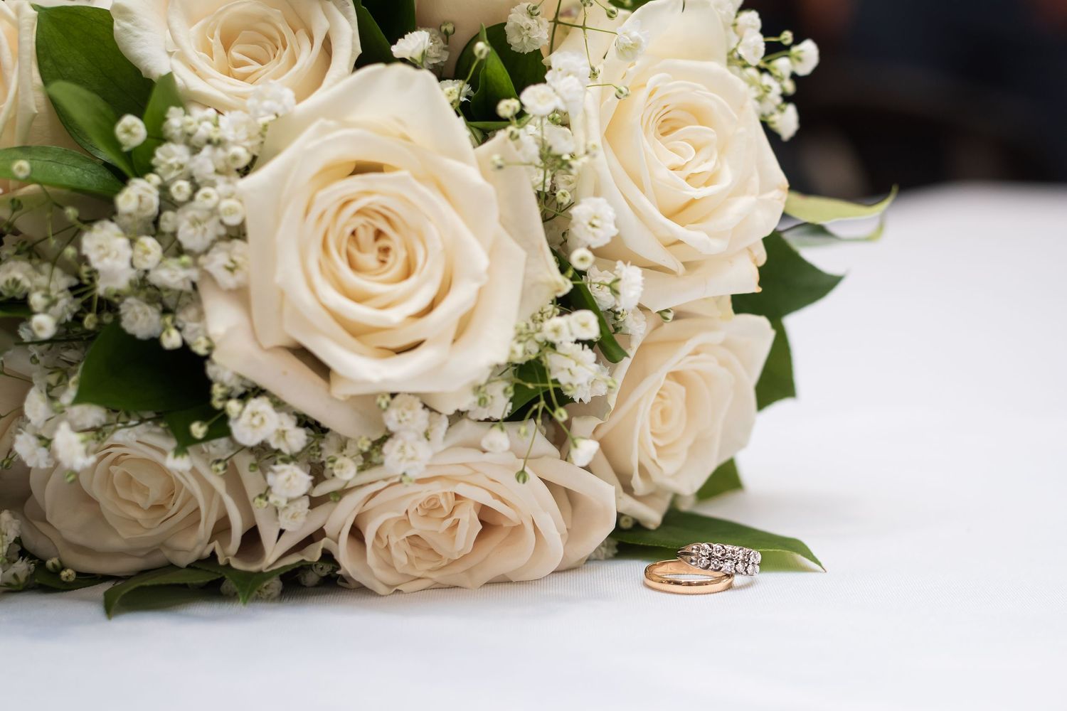 Wedding bouquet of white roses with baby's breath beside two gold wedding rings on a white surface.