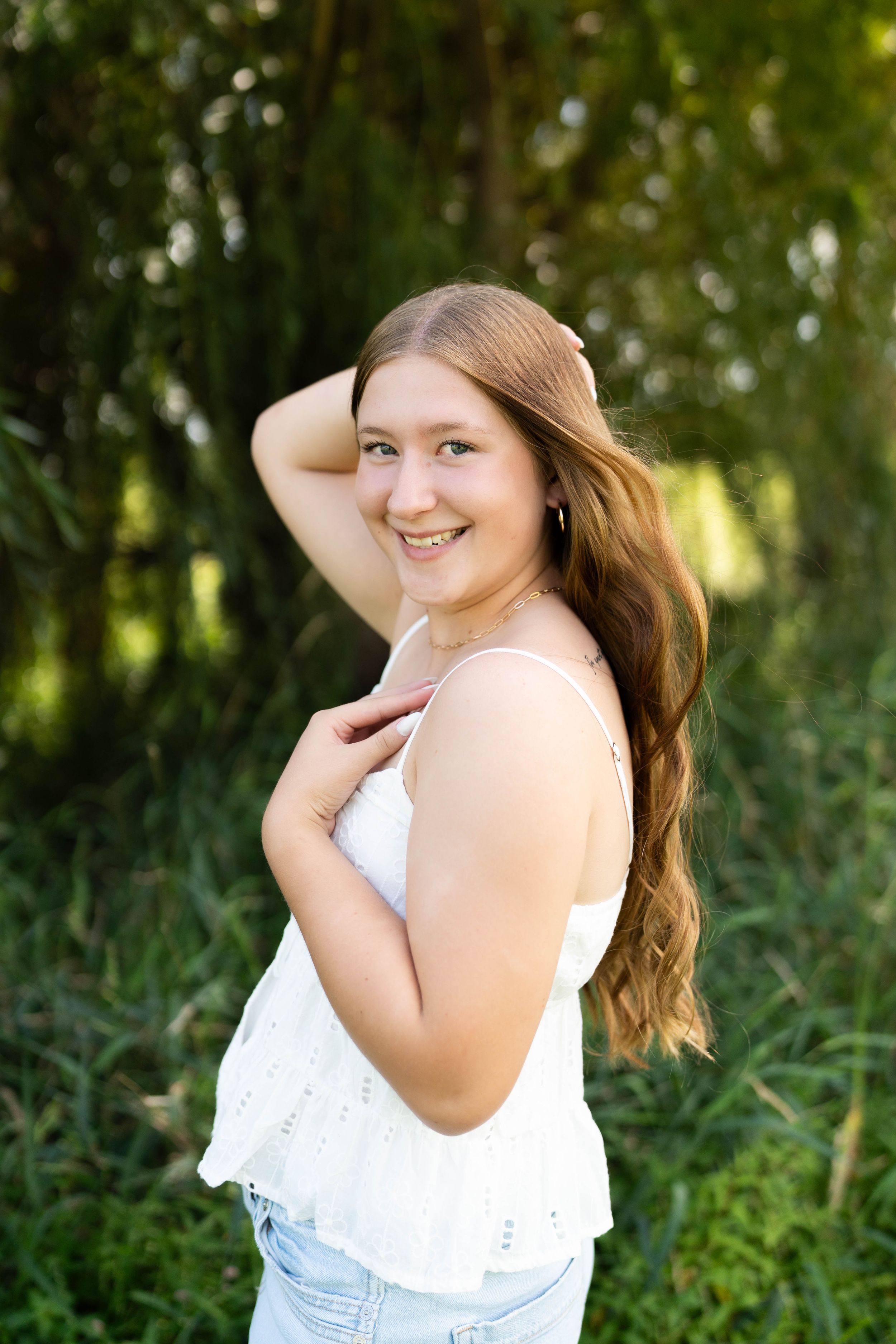 Young woman with long auburn hair smiles outdoors in white top and jeans, posing against lush green foliage.
