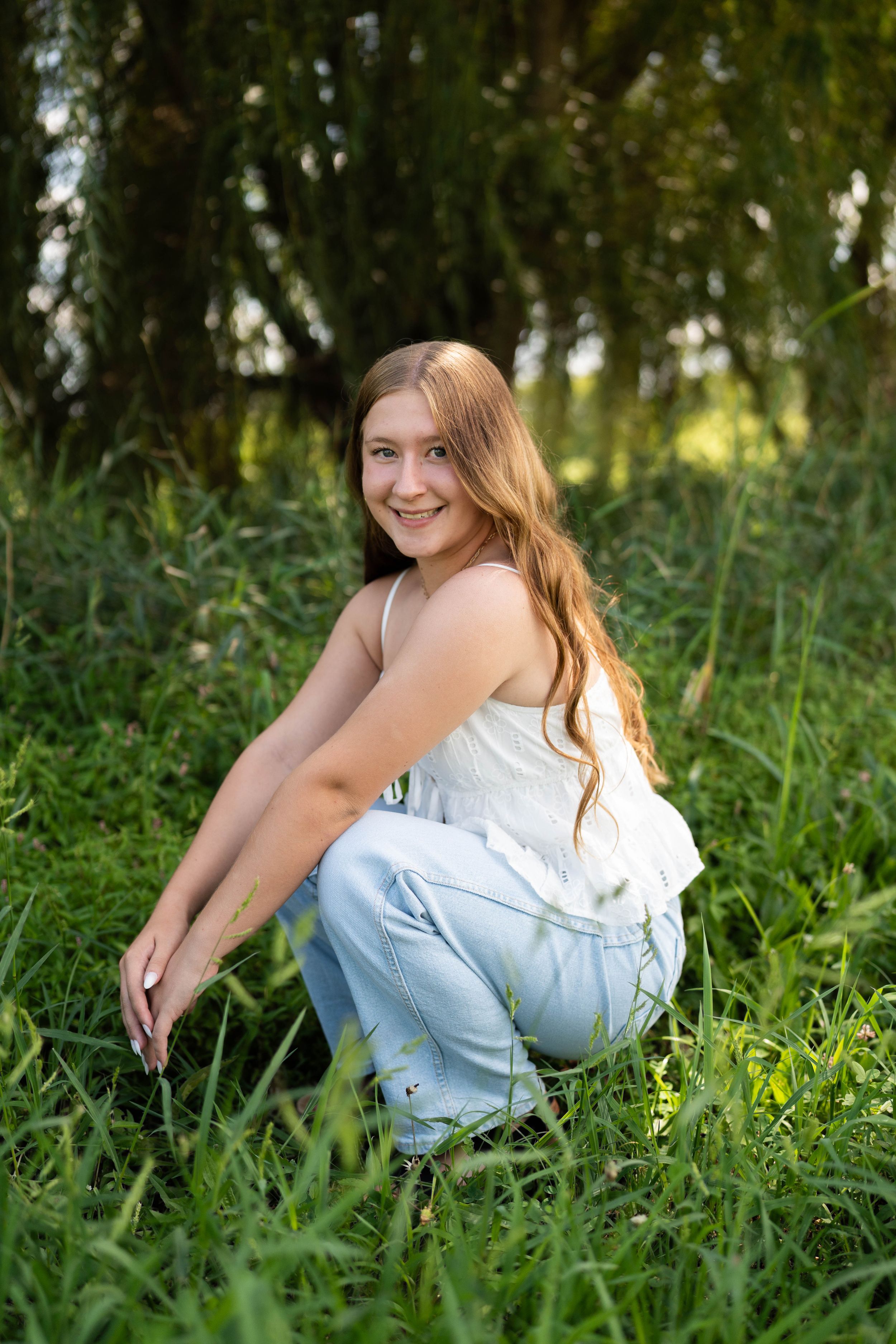 Young woman with long hair crouching in lush green grass, smiling over shoulder, wearing white top and light jeans outdoors.