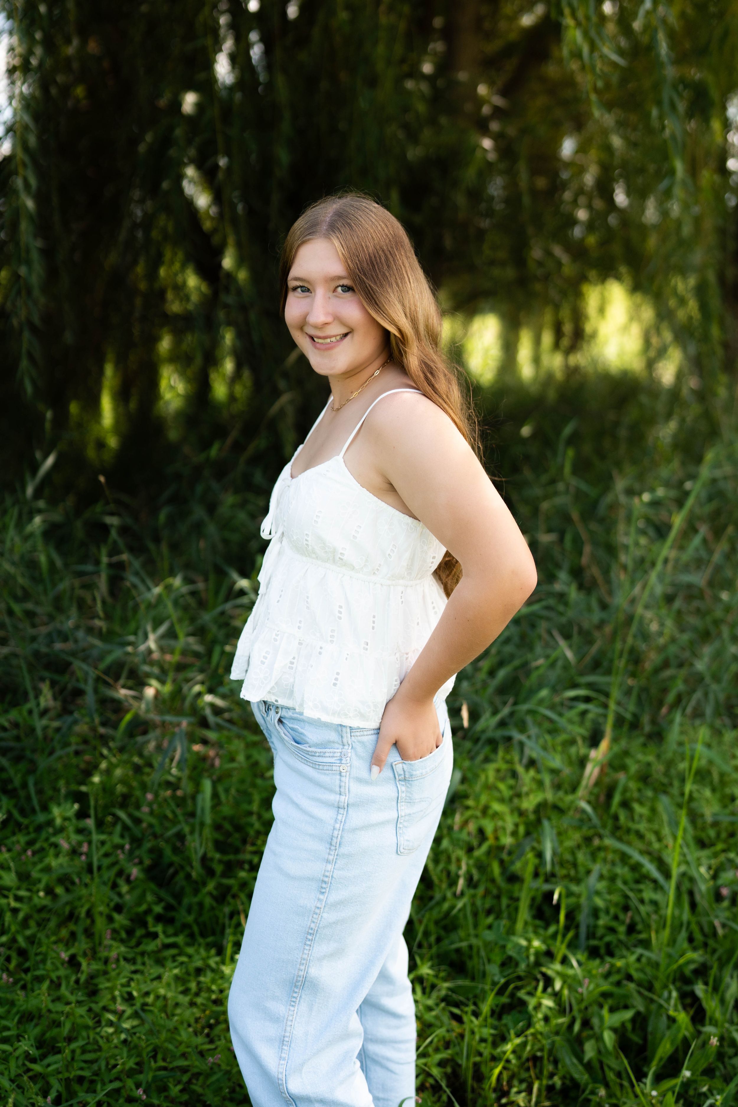 Smiling teenage girl in white ruffle top and light jeans posing outdoors among lush green trees.
