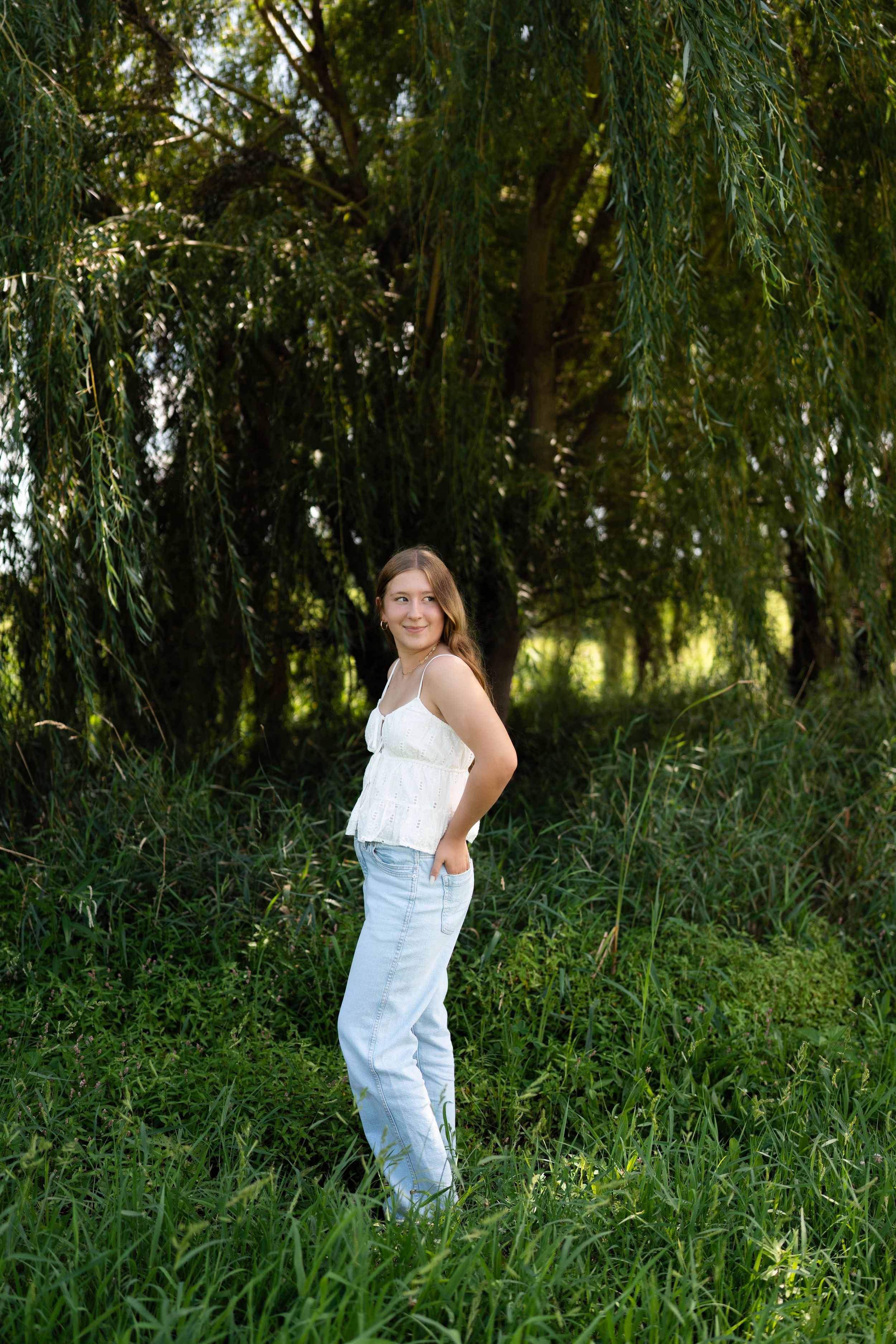 Young woman in white top and light blue jeans posing outdoors among lush green trees and tall grass.