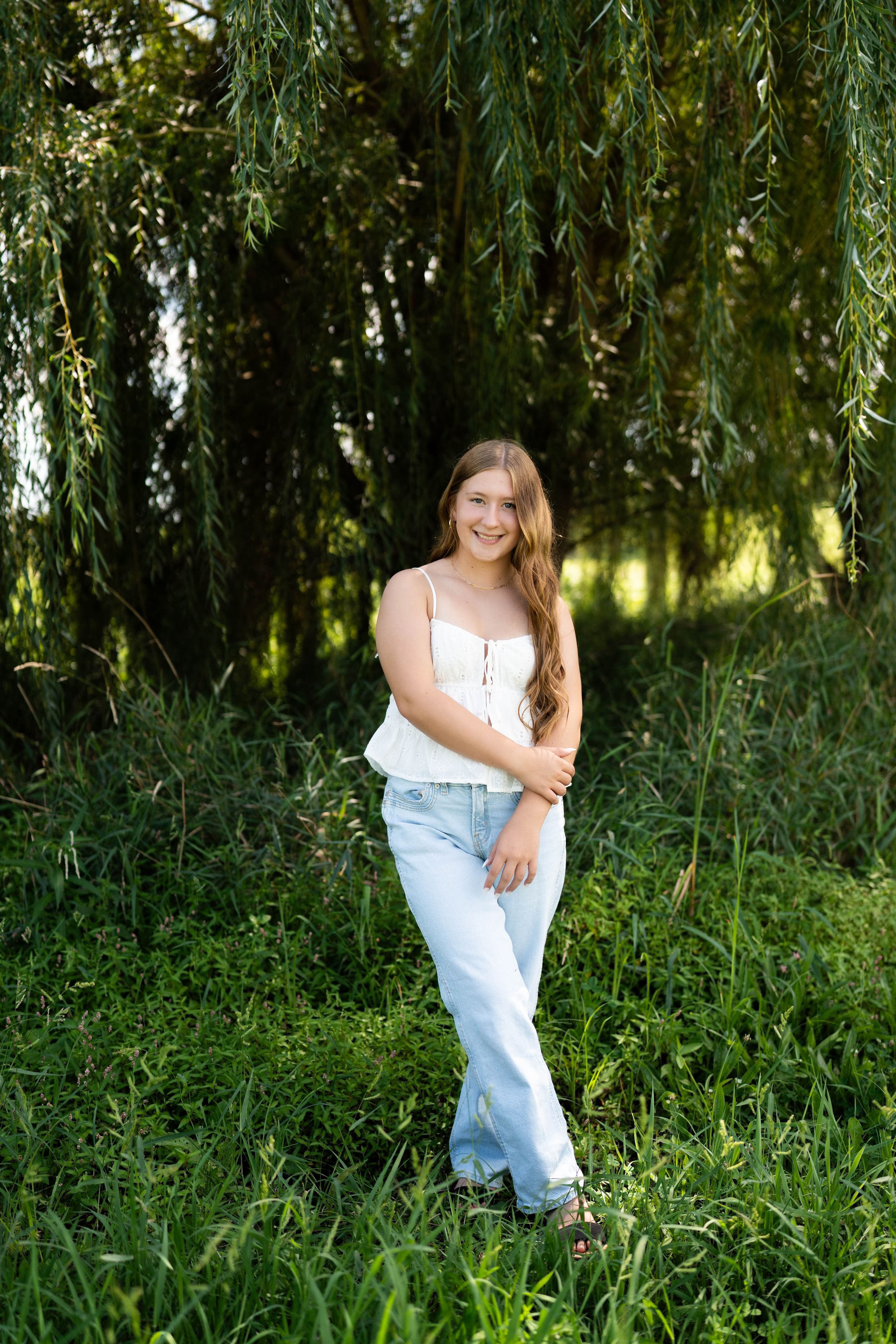 Smiling teenage girl in white tank top and light blue jeans posing outdoors near weeping willow trees.