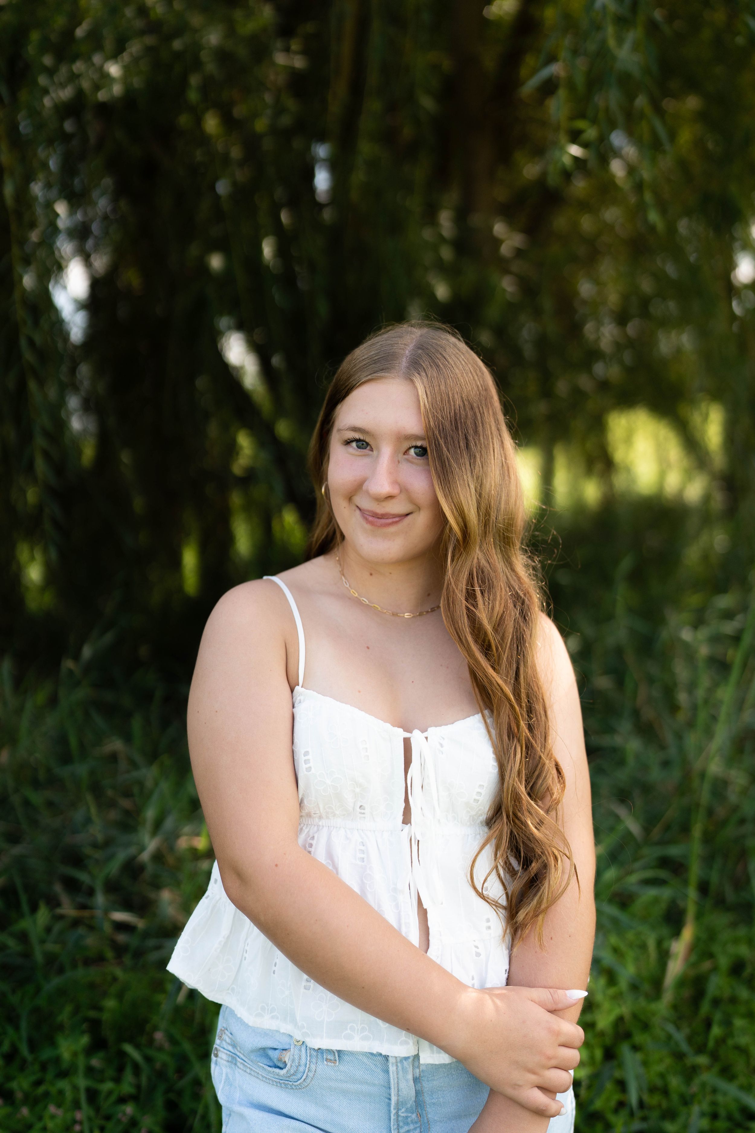 Young woman with long auburn hair in white top and jeans smiling outdoors with lush green trees background.