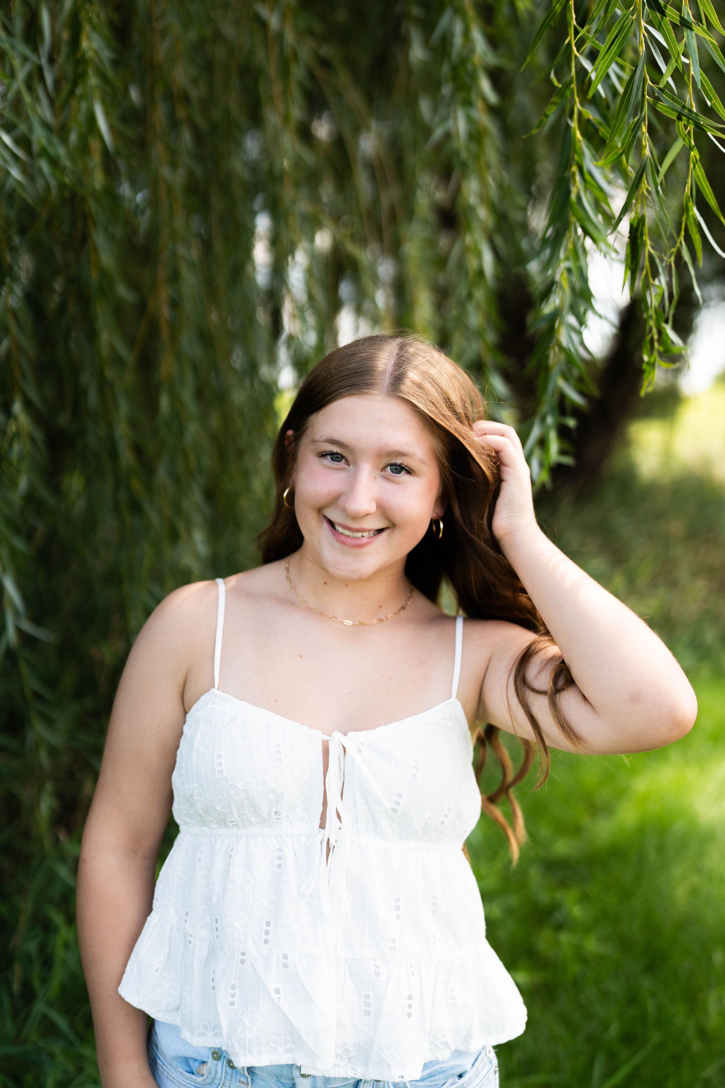 Smiling teenage girl in white eyelet top poses under weeping willow tree, touching her long auburn hair.