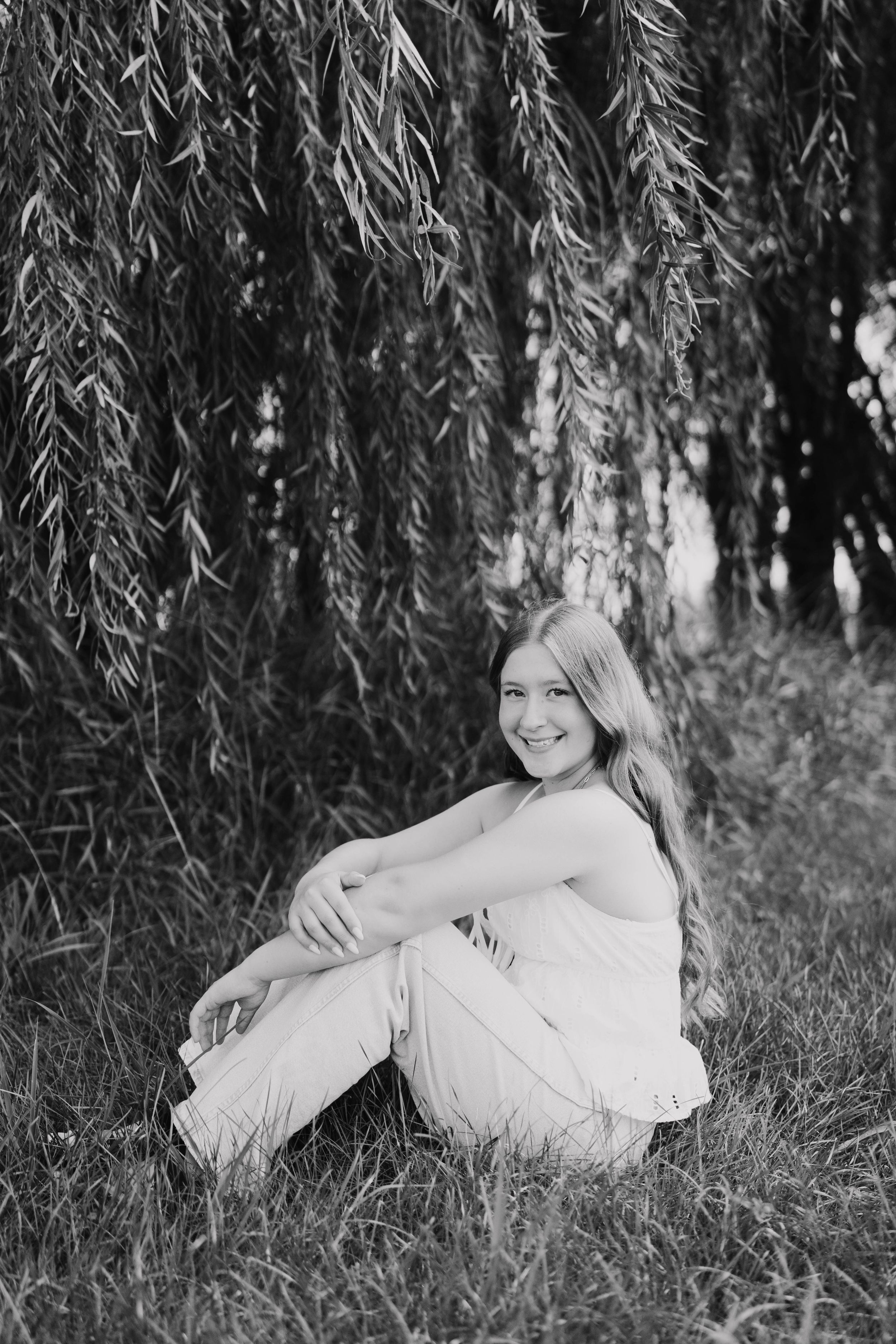 Black and white photo of a smiling teenage girl sitting in grass under a weeping willow tree.