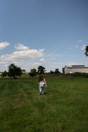 A young girl with curly hair walks through a lush green field toward a farmhouse under a bright blue sky.