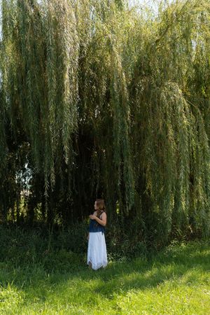 Woman in white skirt standing under a weeping willow tree on green grass.