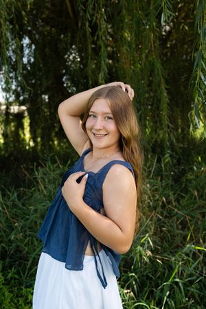 Smiling teenage girl in navy crop top and white skirt posing outdoors near weeping willow tree.
