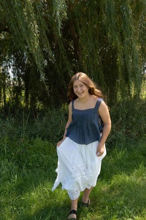 Smiling young woman in navy top and white skirt standing in green grass under a weeping willow tree.