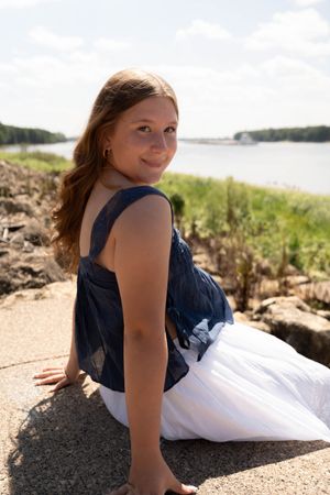 Young woman in navy top and white skirt sitting on stone wall by a river, smiling over her shoulder.