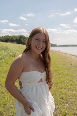 Smiling young woman in white strapless dress standing near a lakeside meadow on a sunny day.