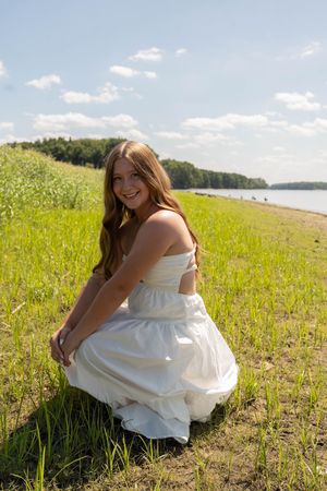 Young woman in white dress crouching in green grass near a lake shore, smiling on a sunny day.
