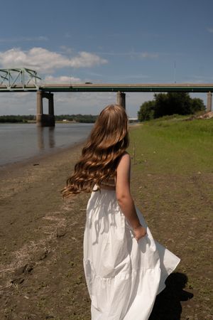 Young woman in white dress walking along riverbank with bridge in background on a sunny day.