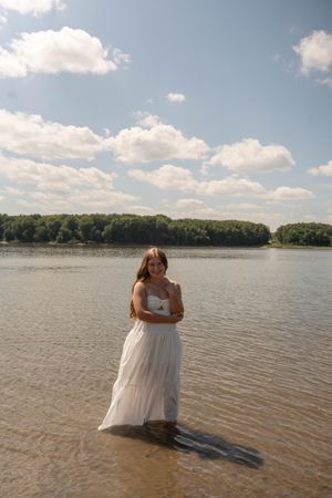 Young woman in white dress standing in a lake with cloudy sky and trees in background.