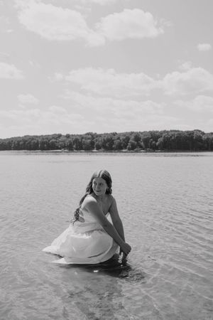 Black and white photo of a bride in a white dress crouching in a lake, with trees and cloudy sky in background.