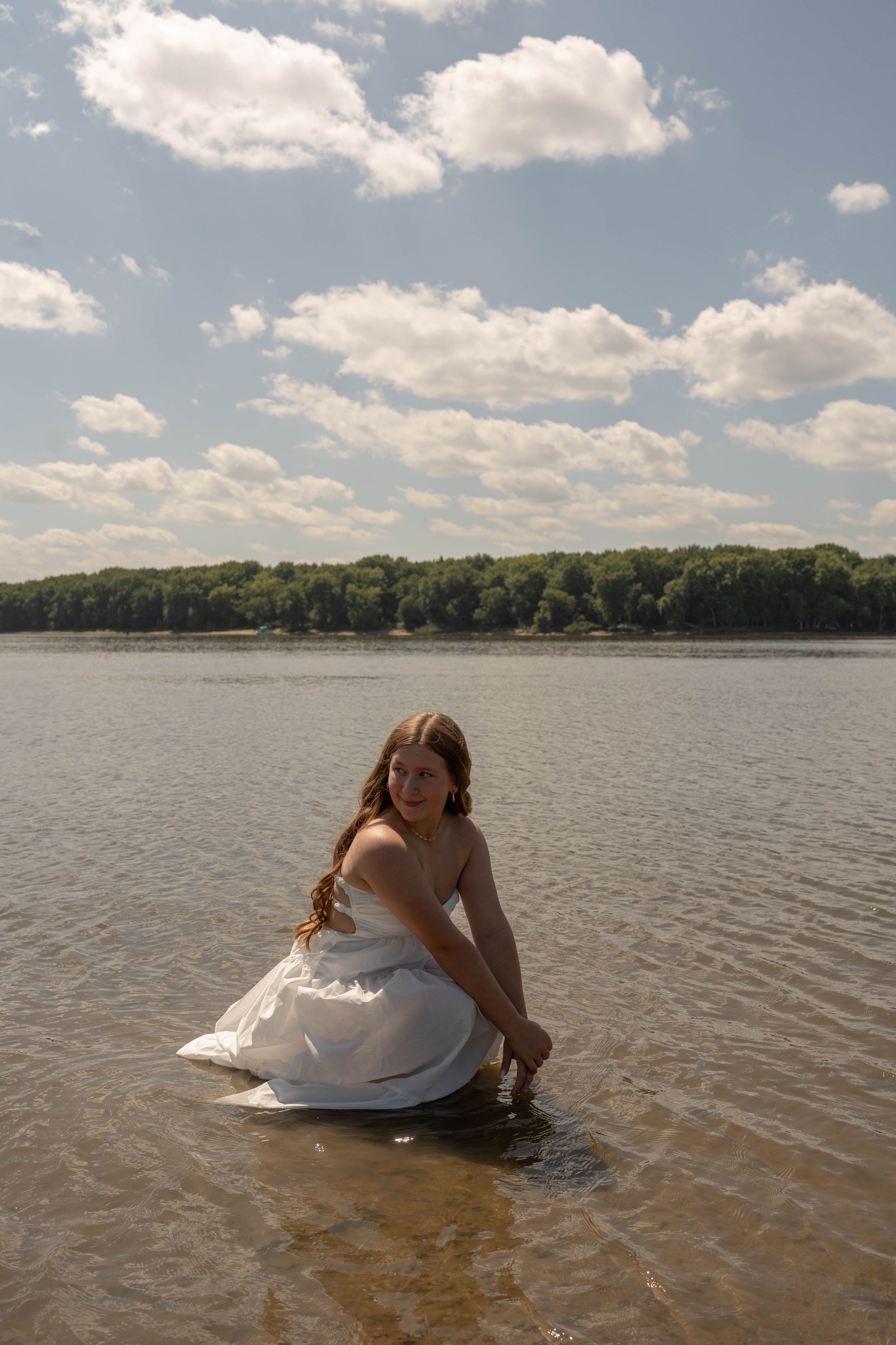 Young woman in white dress crouching in shallow lake water, smiling, with trees and cloudy blue sky in background.