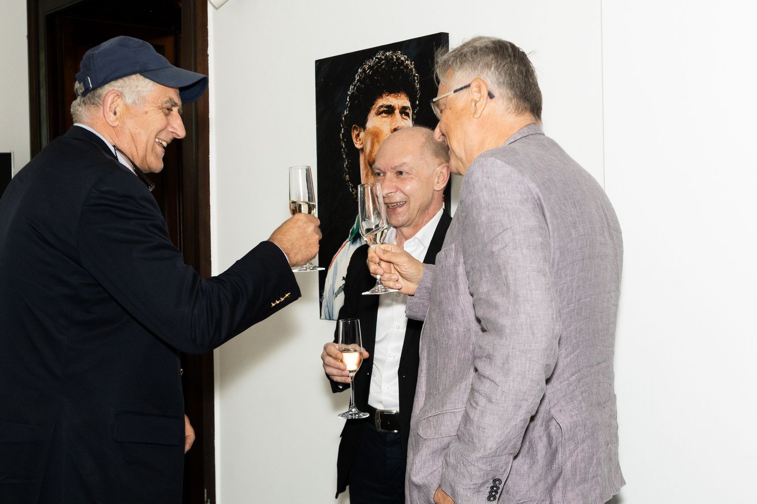 Three men in formal attire toasting with champagne glasses at an art gallery event near a portrait painting.