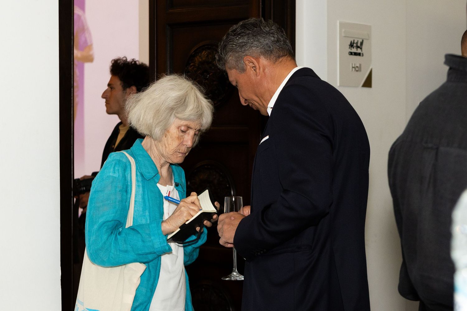 Elderly woman in turquoise cardigan reviewing documents with a suited man holding a wine glass at an indoor event.