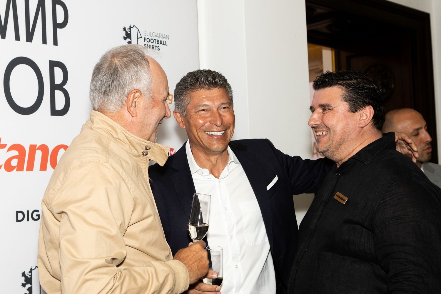 Three men in formal attire laughing and socializing at an upscale event with banners in the background.