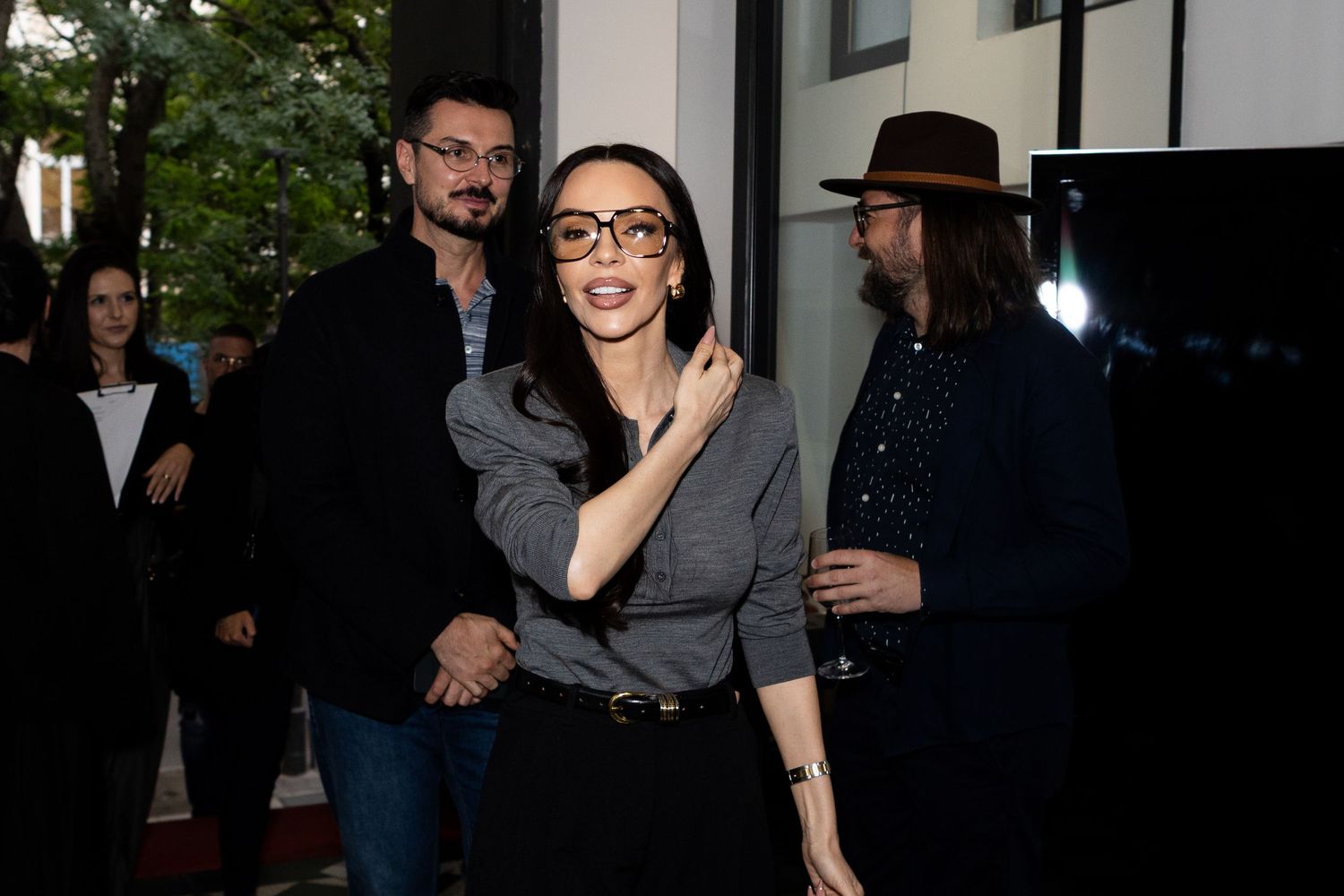 Woman in glasses and gray top poses outdoors with two men at a social event.