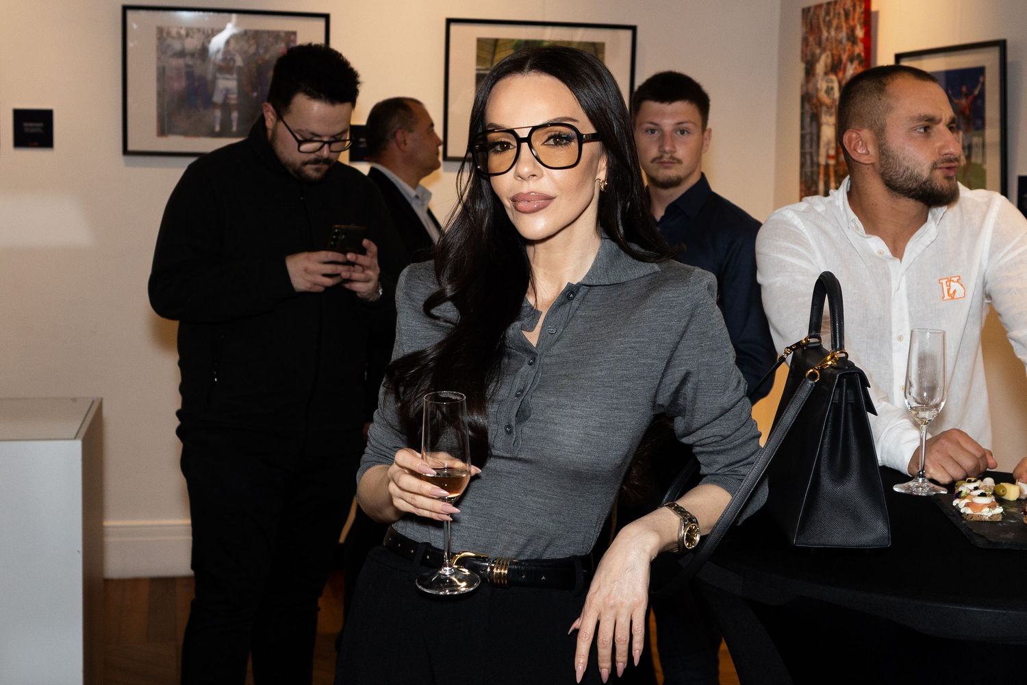 Elegant woman in glasses holding a drink at an art gallery event, wearing a gray top and black pants.