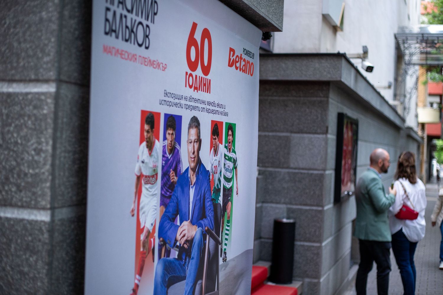 Two people stand near a colorful Betano advertisement poster on a city street corner featuring sports figures.
