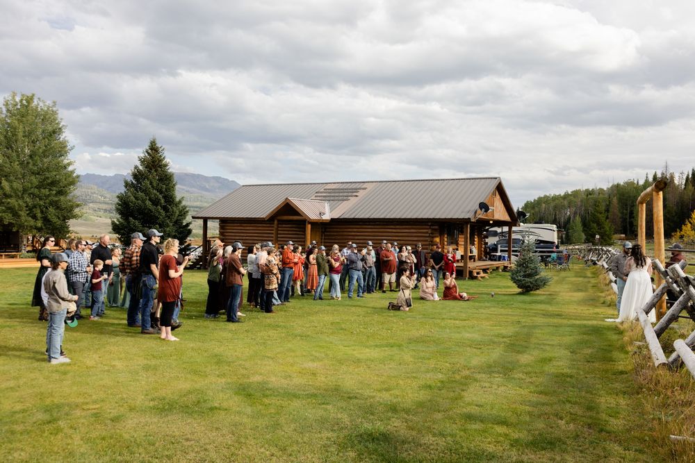 Guests gather on green lawn outside rustic wooden lodge venue surrounded by mountains and trees under cloudy sky.