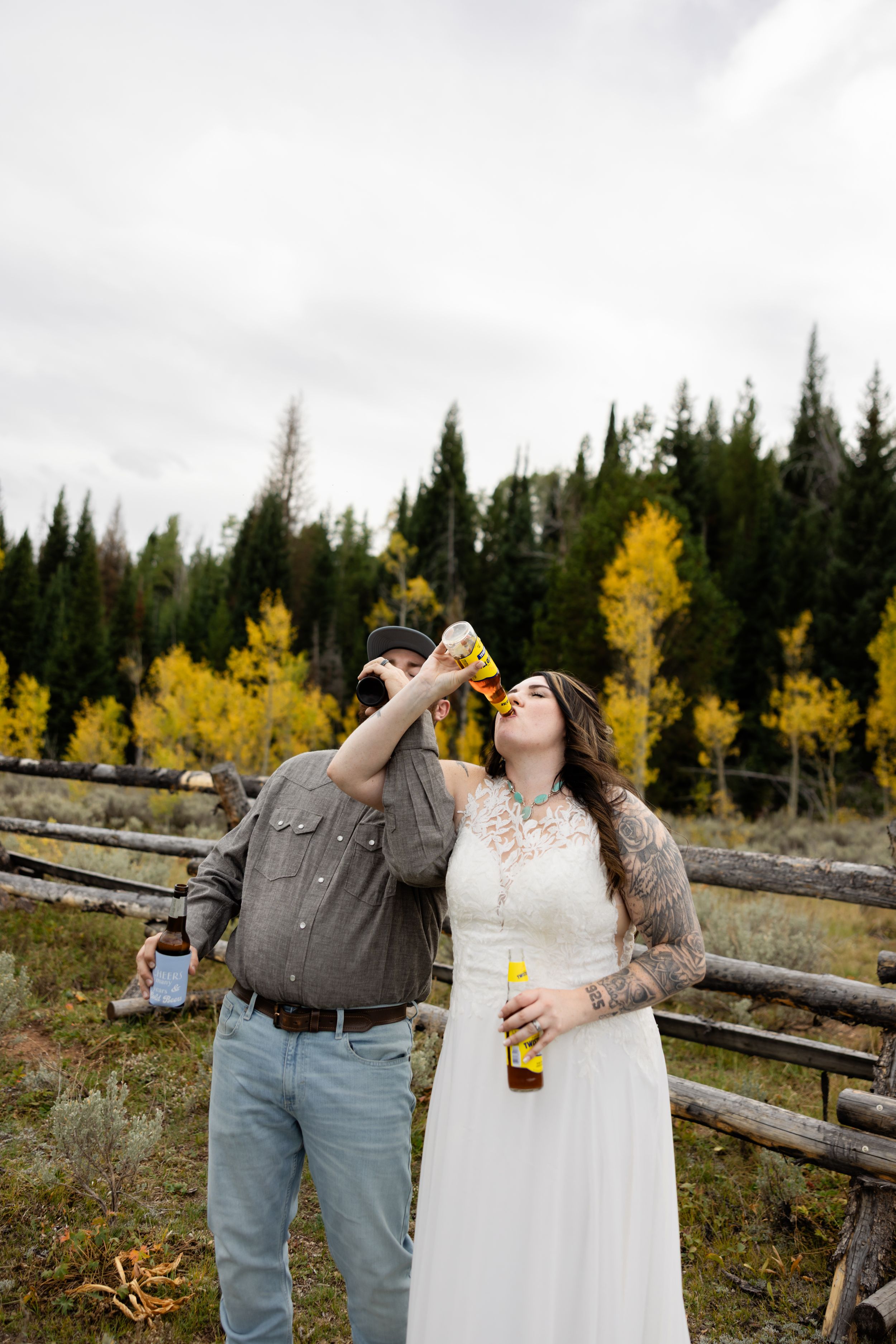 Bride and groom drinking beer bottles outdoors near rustic fence with autumn aspen trees in mountain wedding setting.
