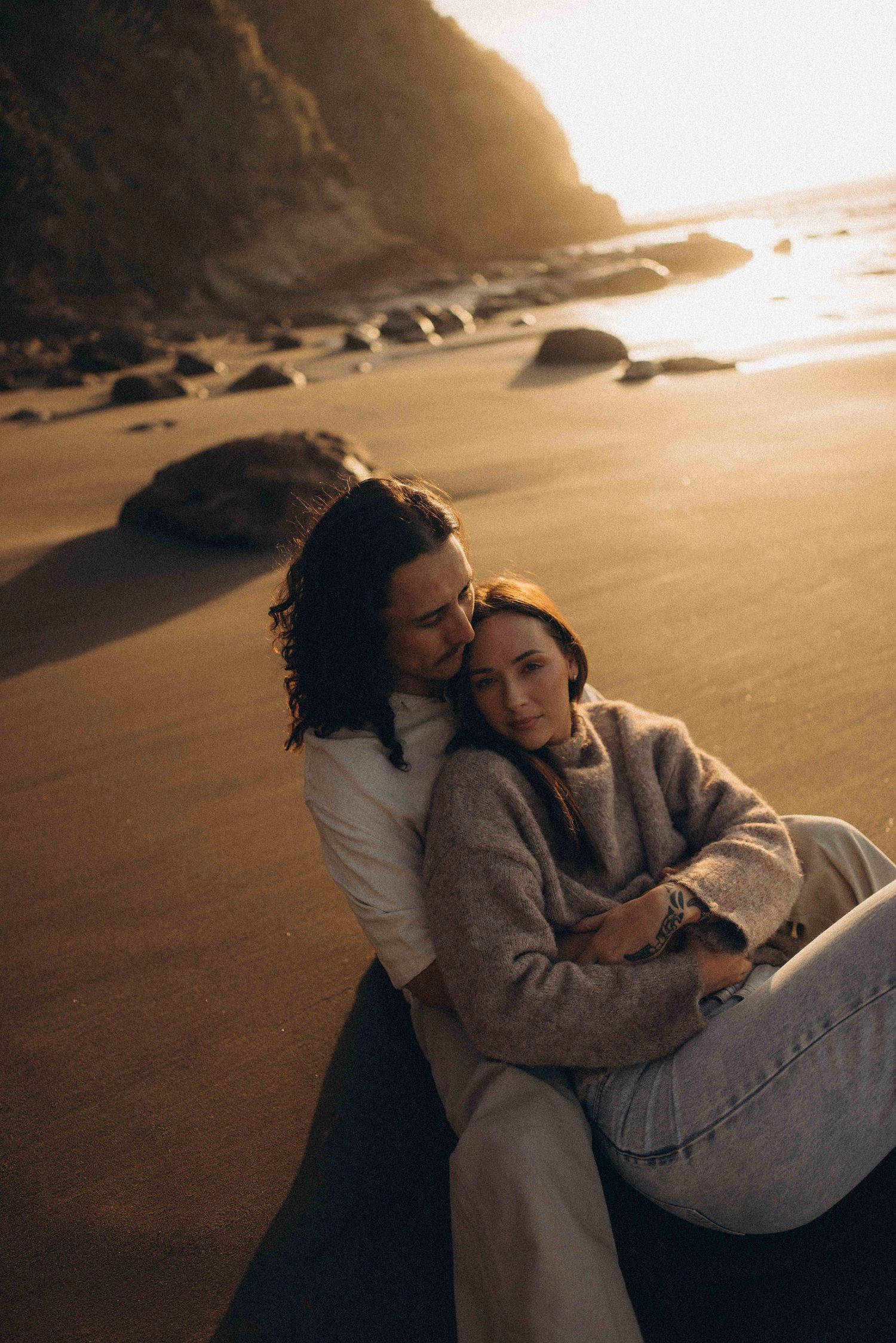 Couple embracing on a sandy beach at golden sunset, sitting together near rocky shoreline with warm glowing light.